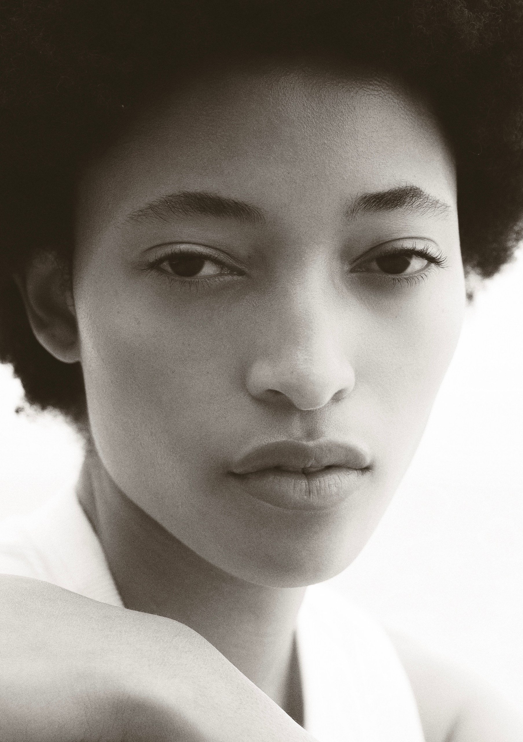 Close-up black and white portrait of a woman with natural curly hair, focused expression, minimal makeup, and soft lighting.