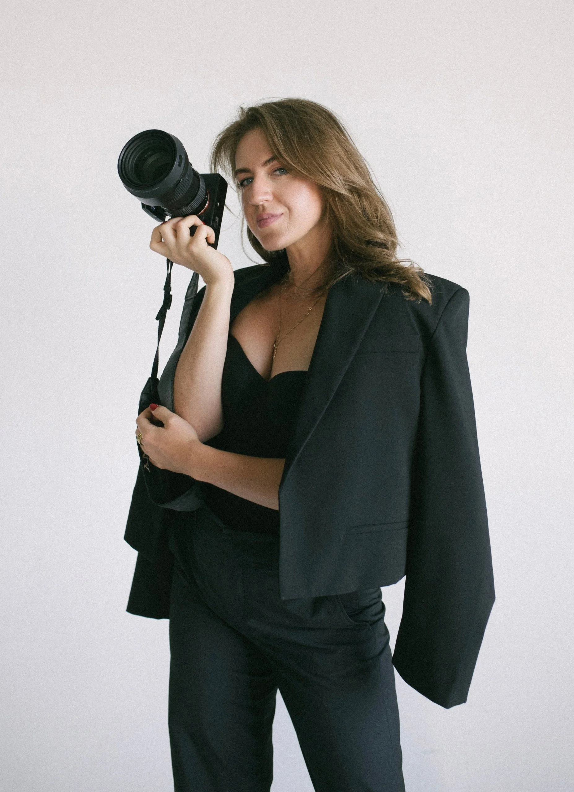 A woman with light brown hair and blue eyes standing against a plain white background, holding a professional camera with a large lens to her face, dressed in a black blazer and matching pants.