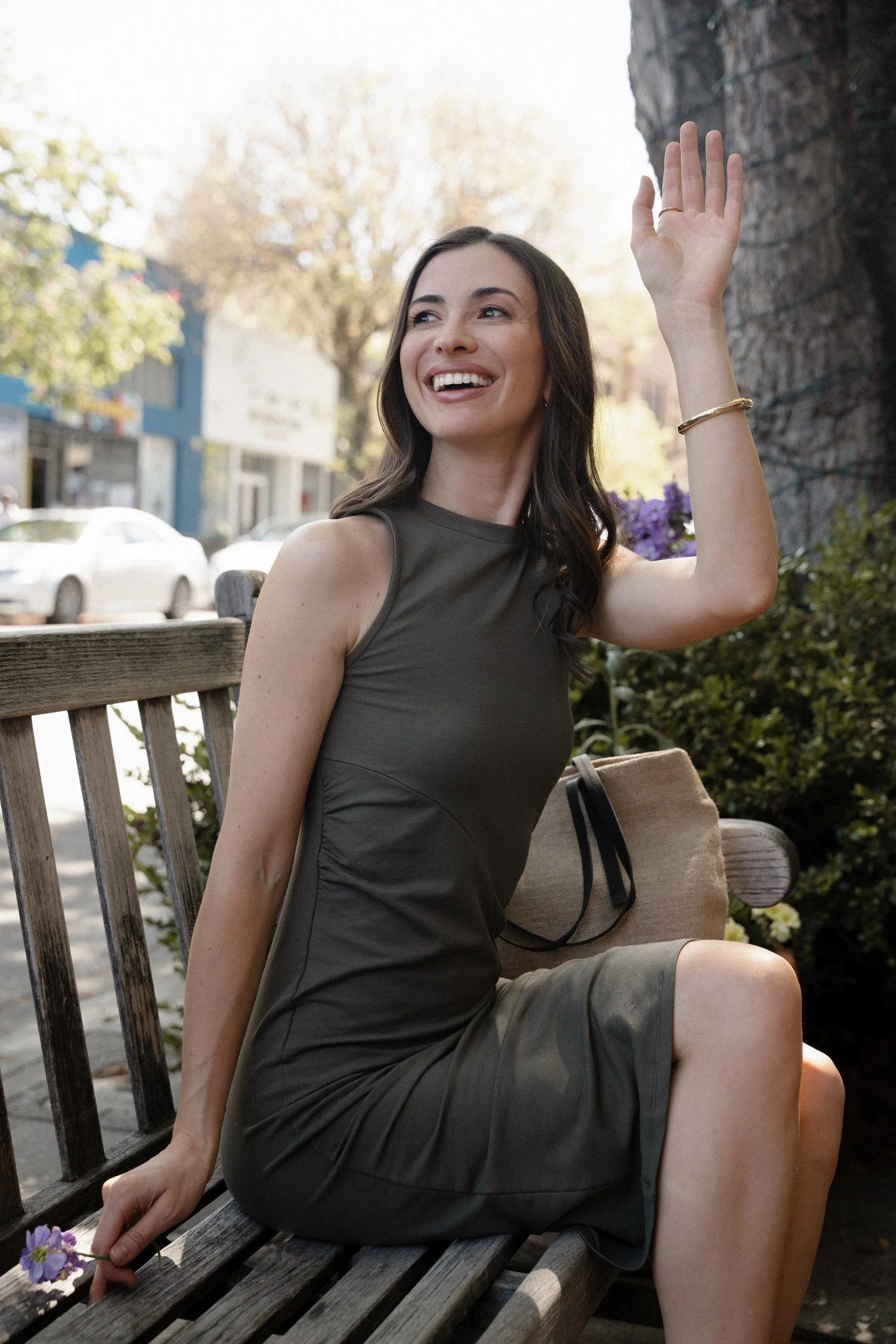 A young woman sitting on a wooden park bench, smiling and waving, with her hand raised and outside in a park with trees, buildings, and cars in the background.
