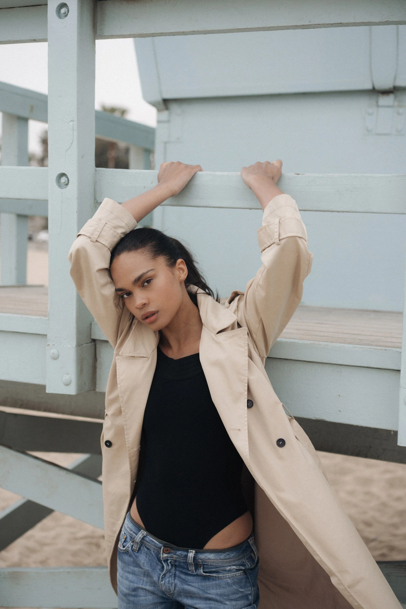 A young woman with dark hair, wearing a beige trench coat, black top, and ripped jeans, leaning against a light-colored wooden structure on a beach.