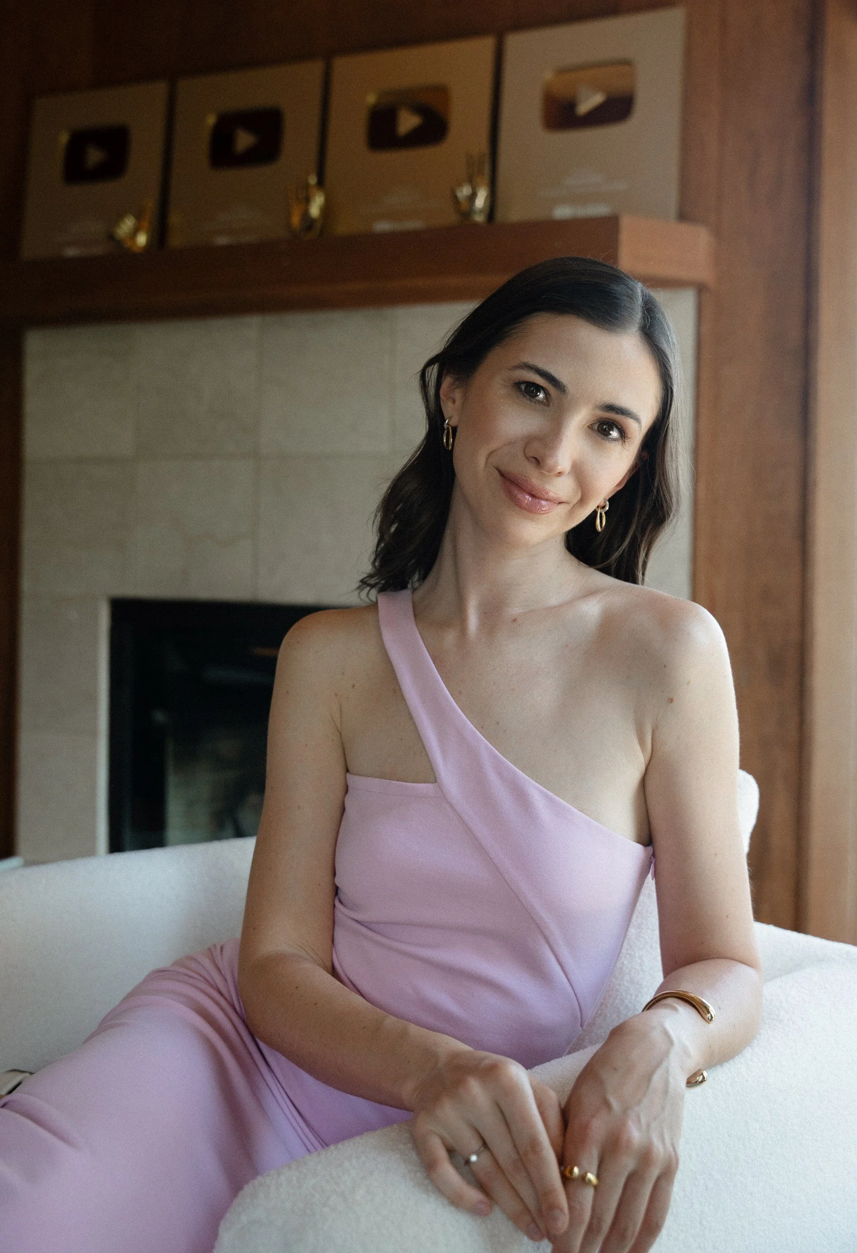 A woman with dark hair and light skin wearing a light purple dress and gold jewelry, sitting on a white chair in a softly lit room with wooden accents and two gold YouTube play button awards in the background.