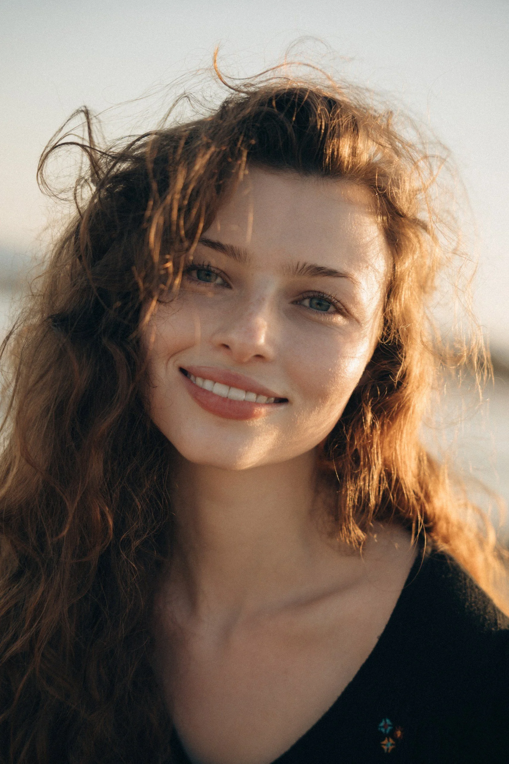 Close-up of a young woman with curly red hair, smiling with white teeth, outdoors during sunset.