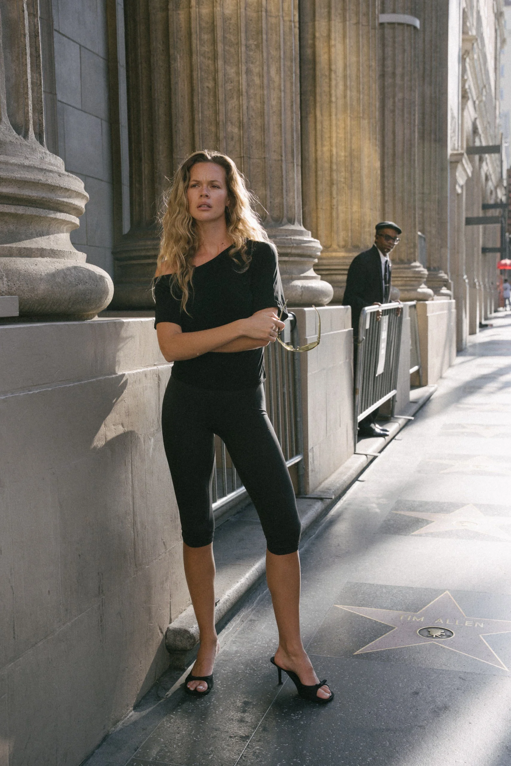A woman in black high heels and black leggings standing on the Hollywood Walk of Fame with her arms crossed, holding sunglasses, in front of buildings with large columns. A man is in the background near a metal barricade.