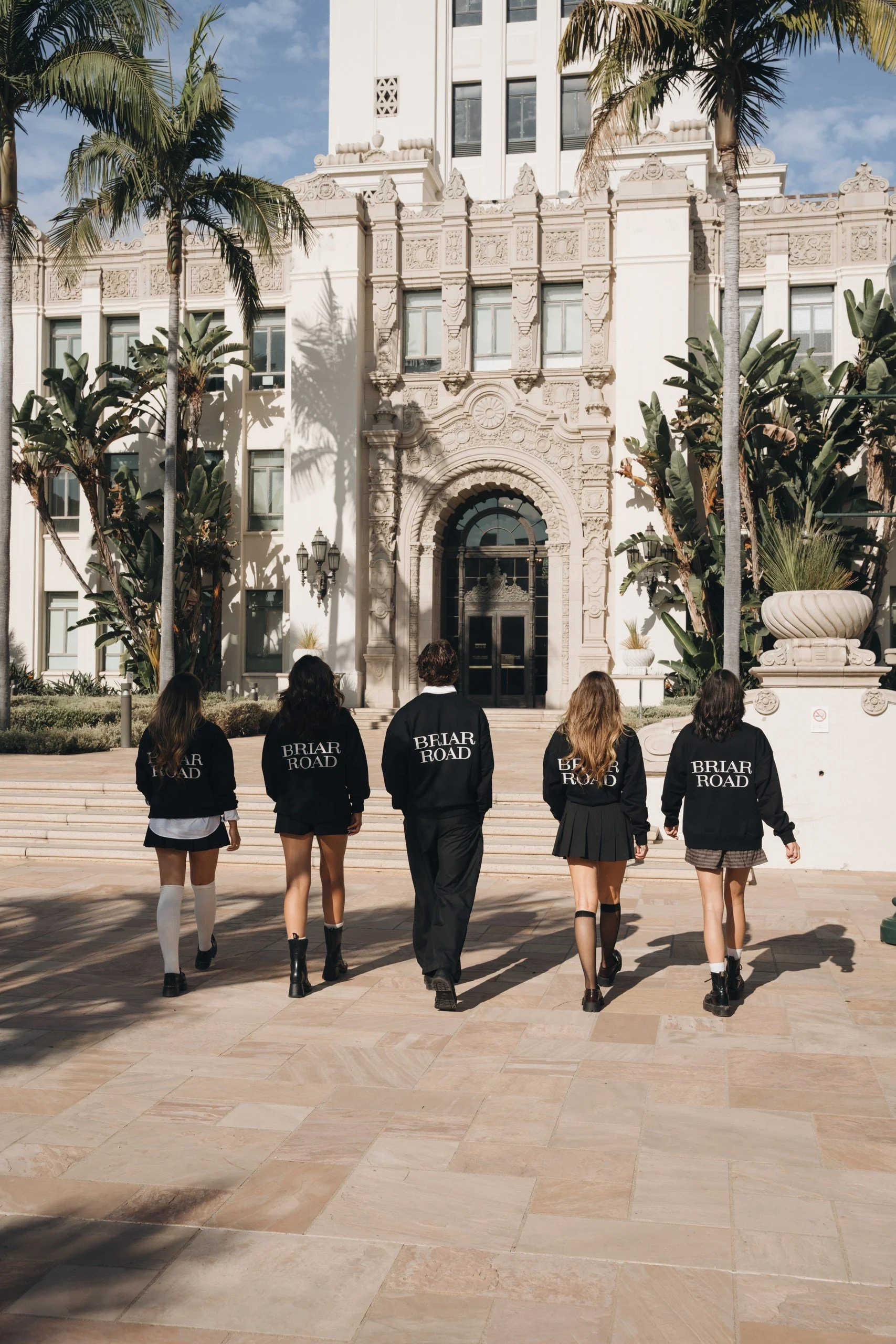 A group of five students wearing Briar Road school uniforms walk towards a historic building with ornate architecture and palm trees. The students are wearing black jackets with the school's name on the back, skirts or pants, and boots.