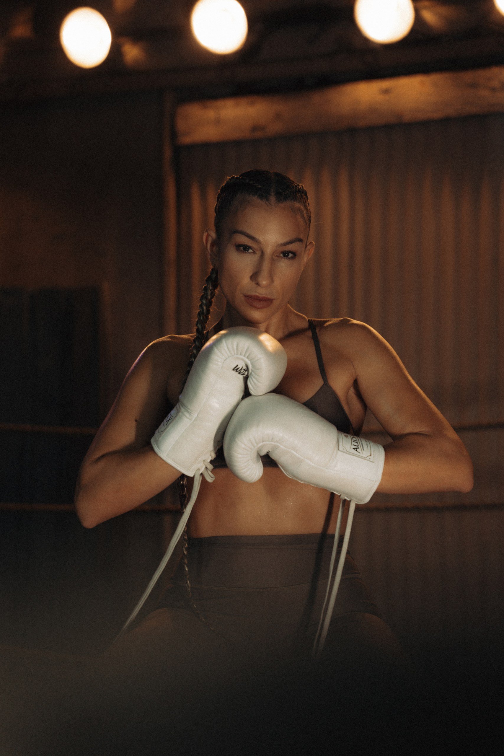 A woman with braided hair wearing boxing gloves and sports bra, posing in a dimly lit gym.