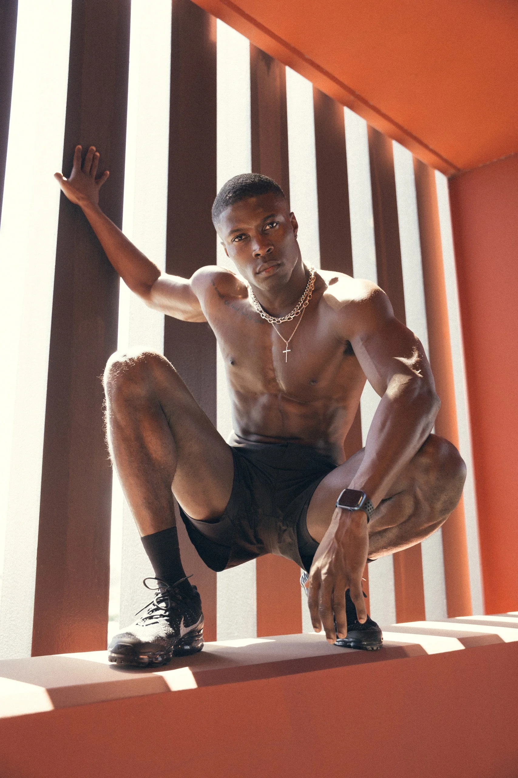 A young man wearing black shorts, running shoes, and a watch is performing a one-arm plank pose on a ledge with vertical orange and brown architectural panels in the background. He is shirtless, has tattoos on his arm and shoulder, and is wearing lay