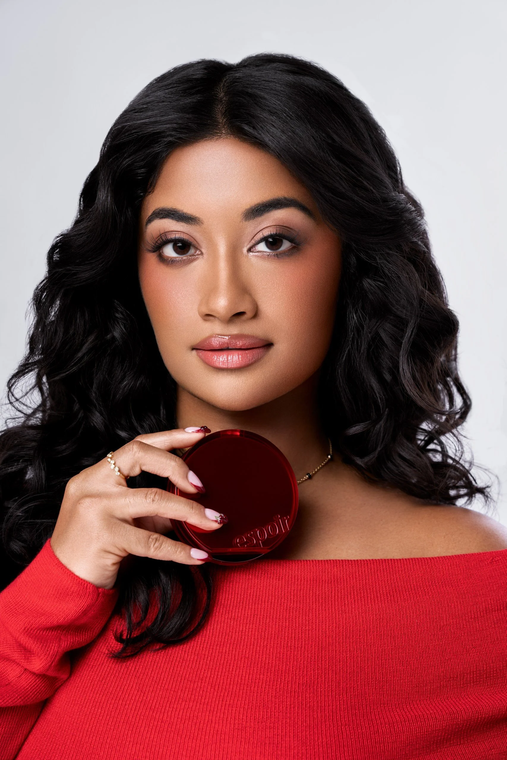Close-up of a woman with long black wavy hair, holding a red compact makeup mirror, wearing a red top, with makeup and manicured nails.