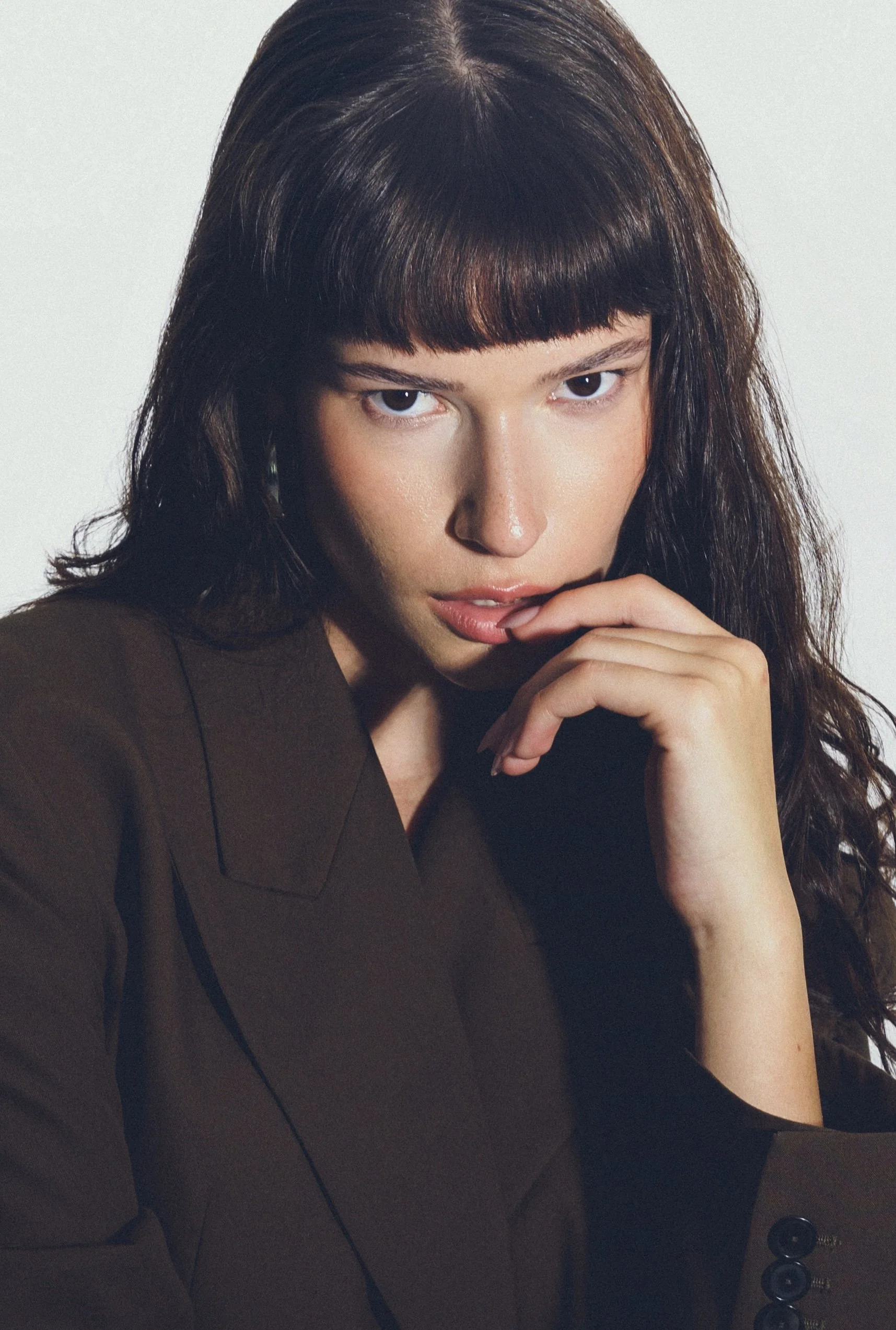 A woman with dark brown hair and bangs, wearing a dark blazer, looking directly at the camera with her hand near her mouth.