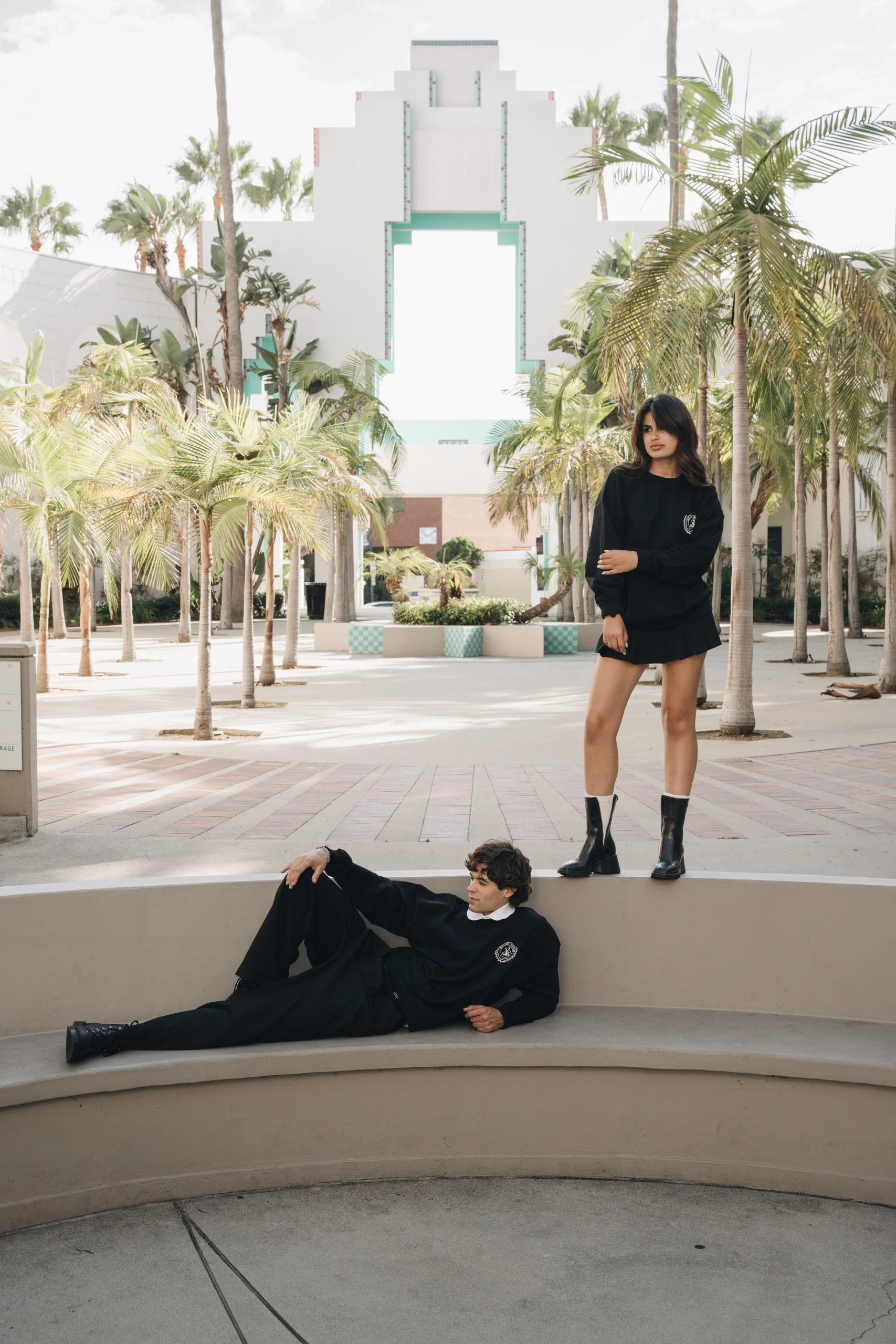 A young woman in a black sweatshirt and skirt standing next to a young man in black sitting on a ledge in an outdoor setting with palm trees, a white building in the background, and bright sky.
