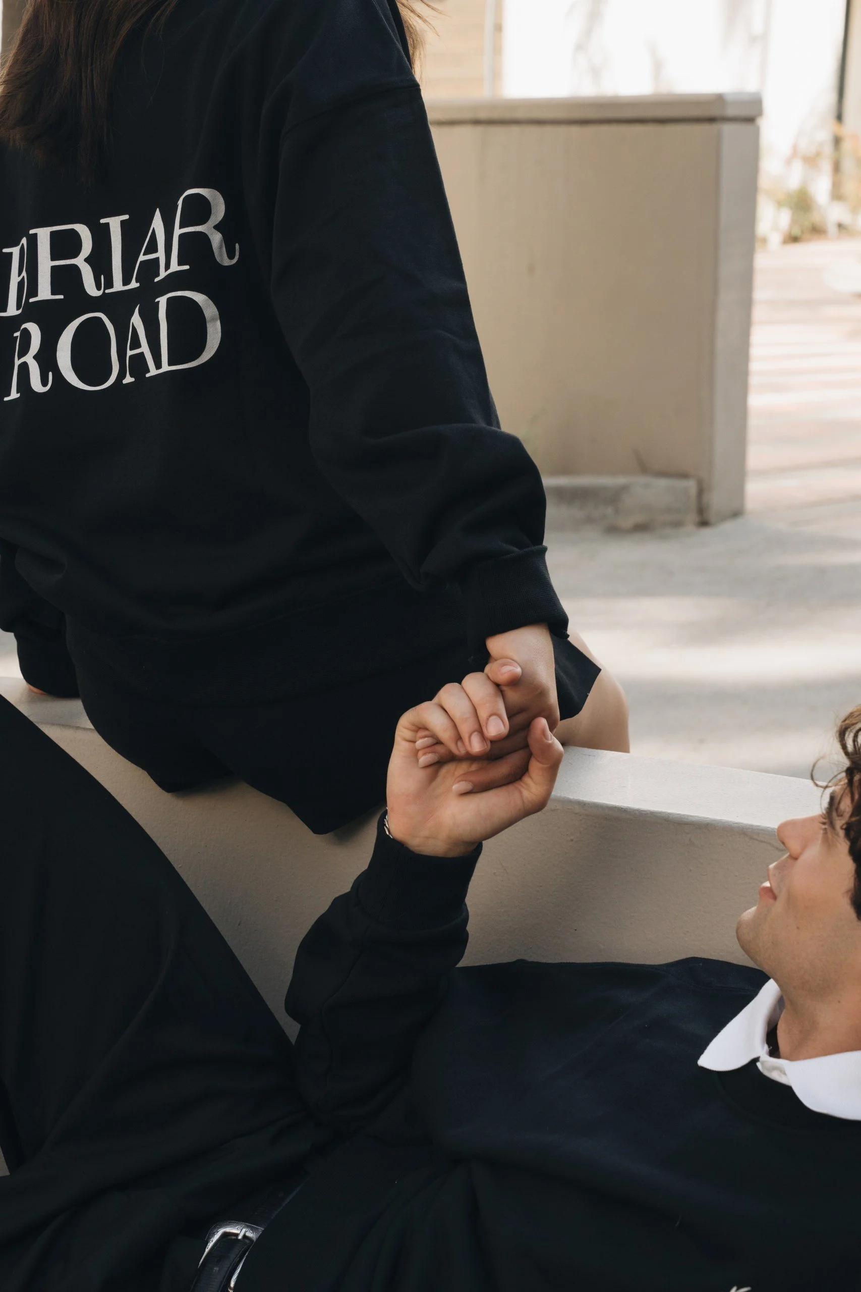 A person wearing a black jacket with white text that says 'BIRRIA ROAD' is sitting at a table. Another person with a white shirt is sitting across from them, and they are holding hands.