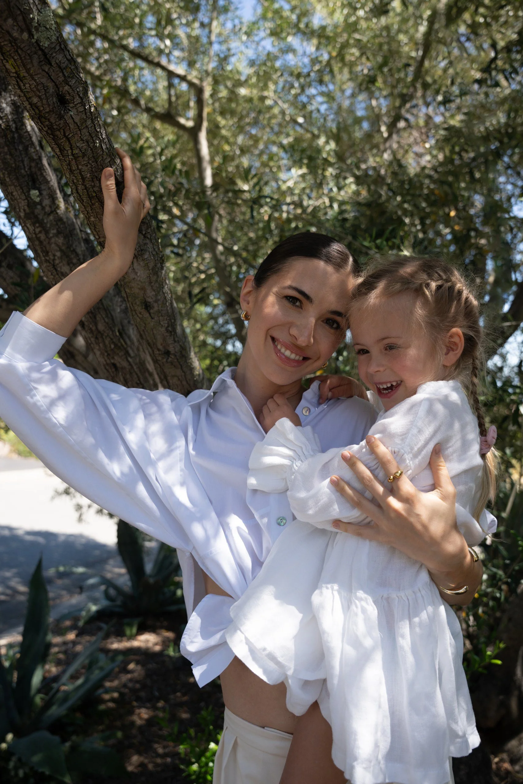 A woman and a young girl smiling and hugging outdoors near a tree, wearing white clothing, with greenery and sunlight in the background.