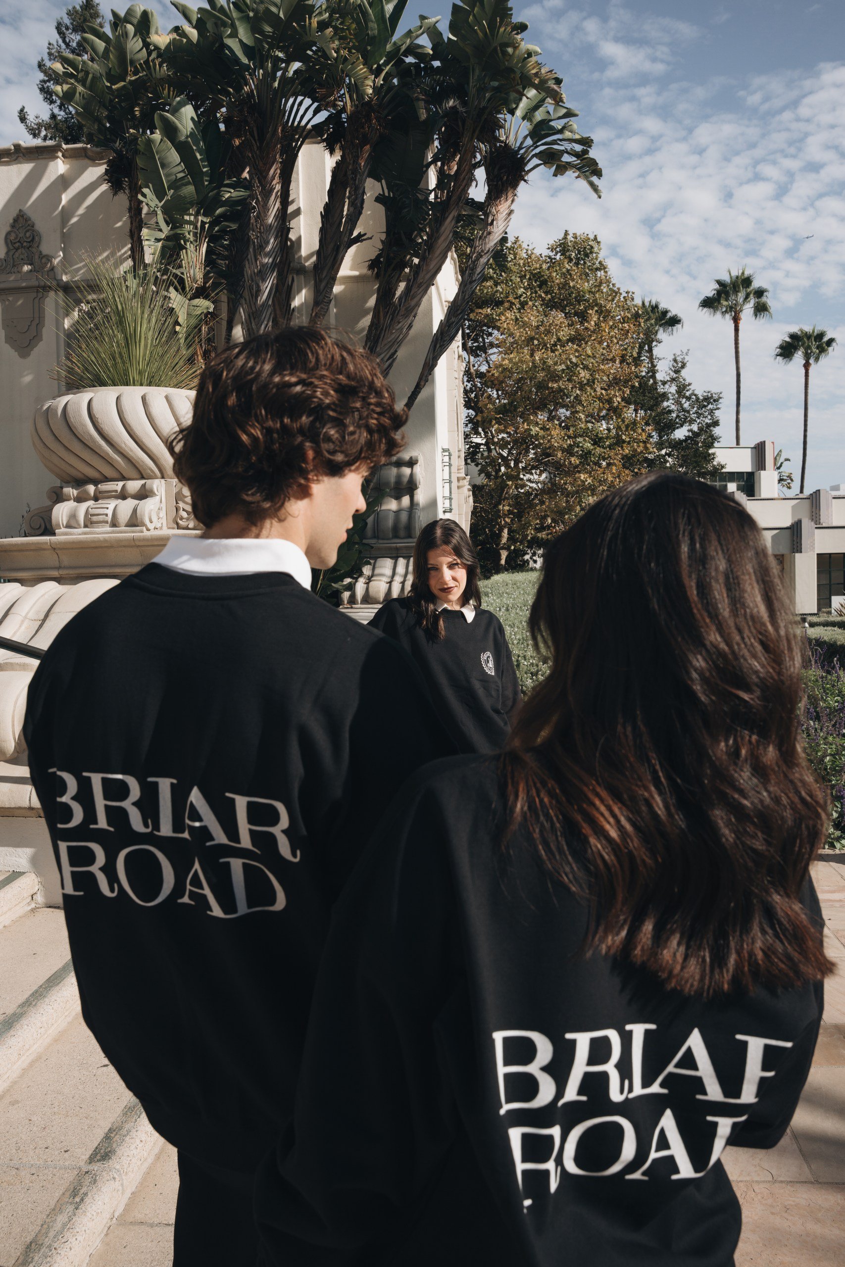 Group of people standing outdoors near a building with trees and a partly cloudy sky in the background. Some individuals wear black sweatshirts with the words 'BRIAR ROAD' printed on the back.