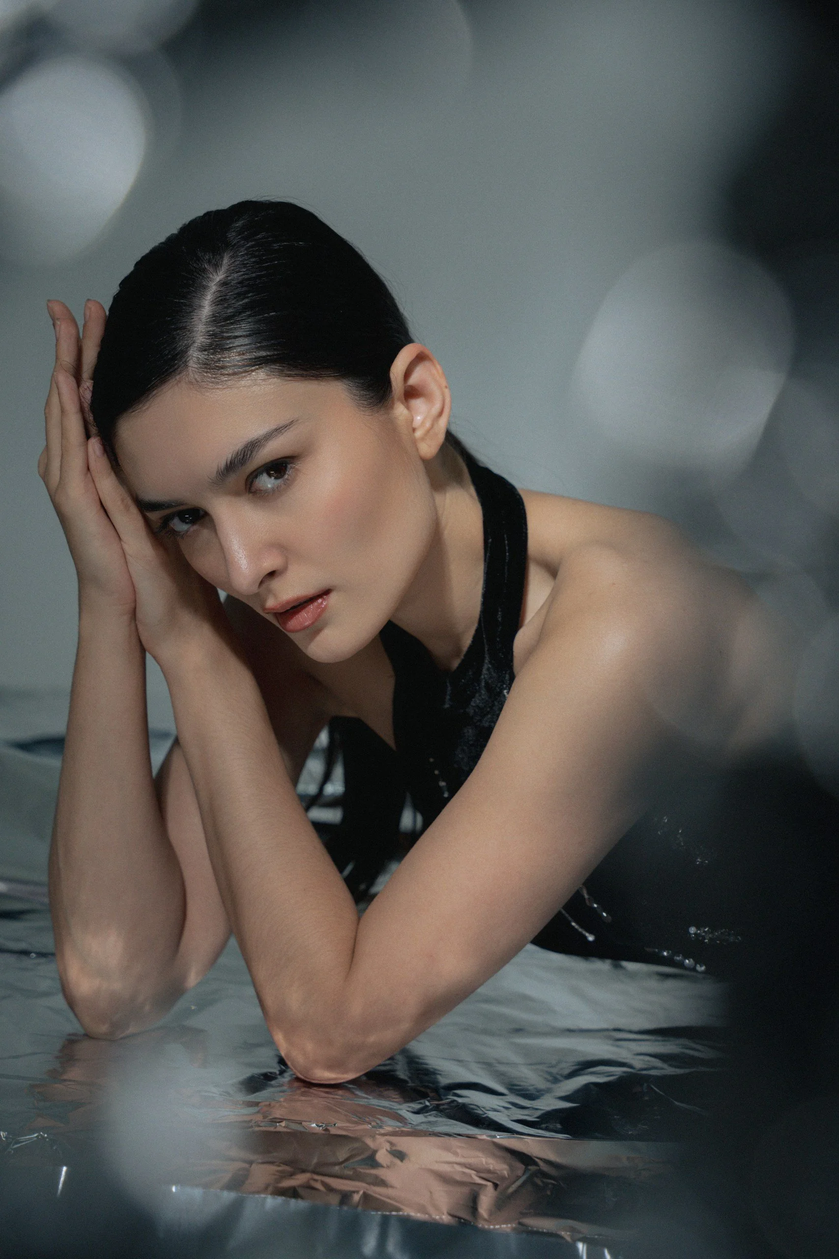 A young woman with dark hair styled in a sleek look, resting her head on her hands with her elbows on a reflective surface. She is wearing a sleeveless black top with a shiny fabric, and the background is blurred with circular light reflections.