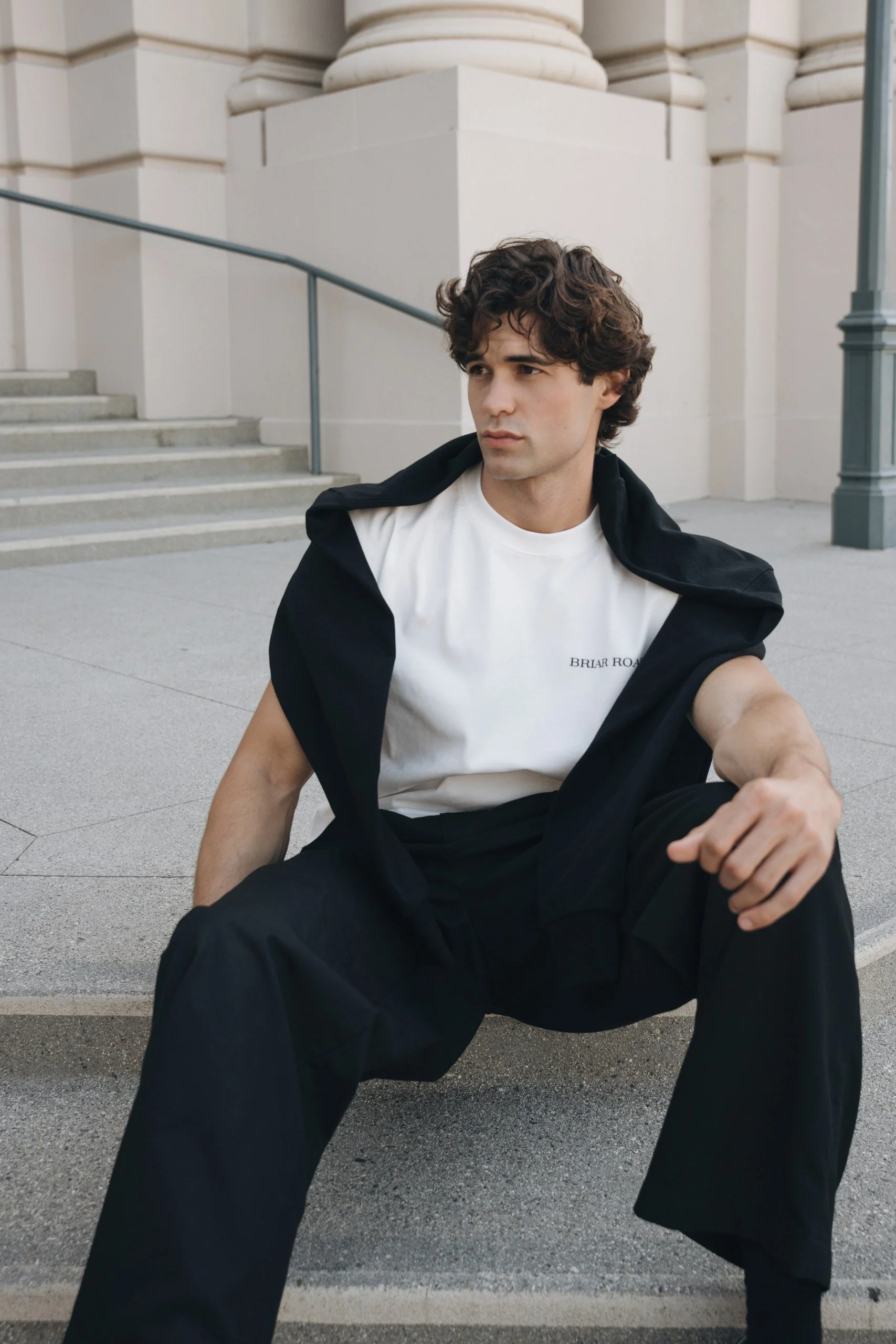 A young man with curly hair wearing a white t-shirt and black pants, sitting on a curb outside a white building with stairs and a railing, looking off into the distance.