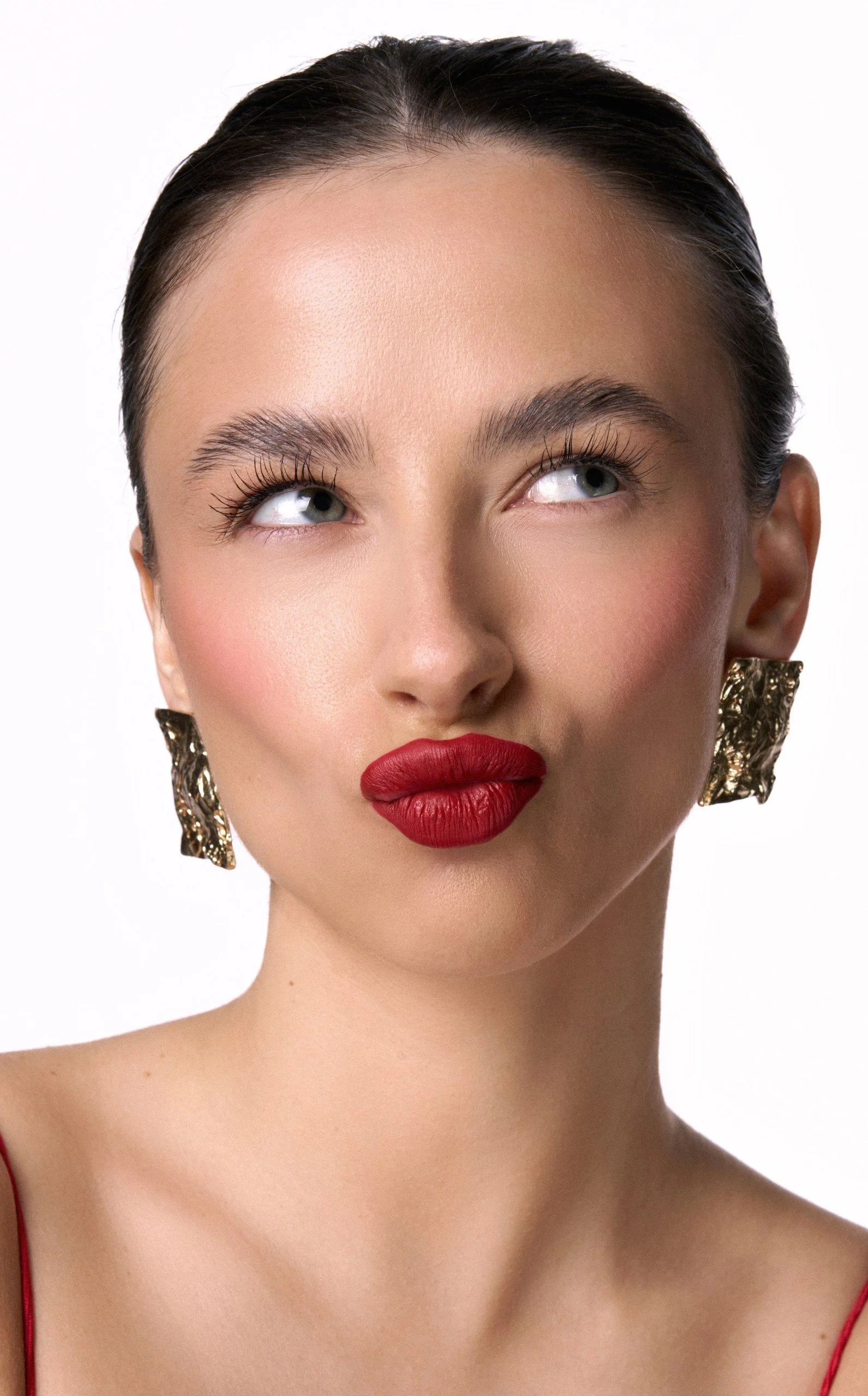 Close-up of a woman with dark hair pulled back, wearing bold red lipstick, eye makeup, gold statement earrings, and a red strap garment.