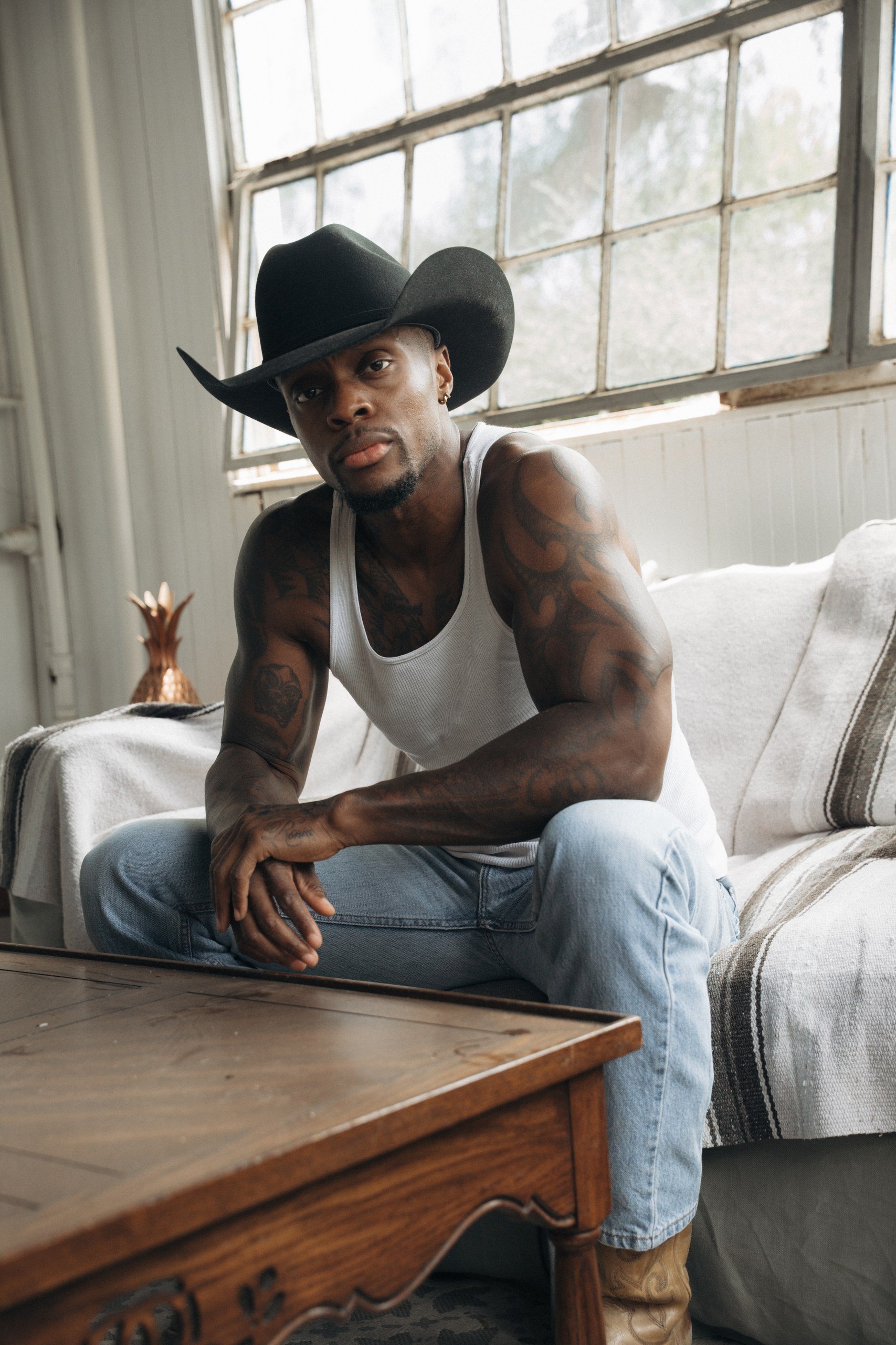 A young man with tattoos and a beard is sitting on a sofa. He is wearing a white tank top, light blue jeans, and a large black cowboy hat. The room has a large, multi-pane window in the background and a wooden table in the foreground.