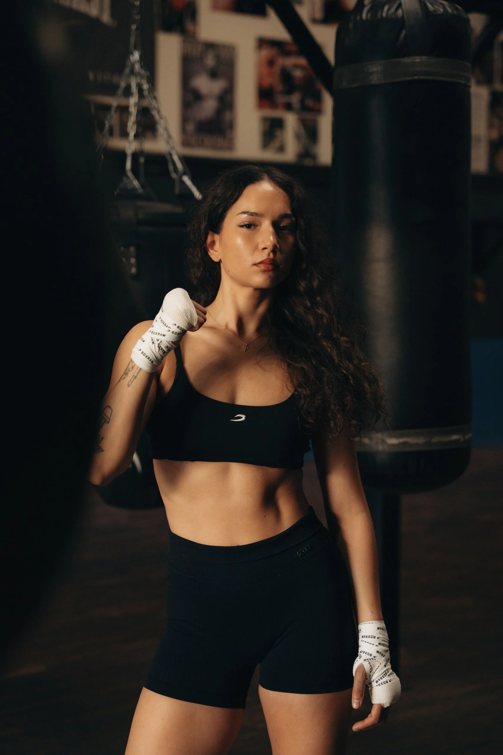 A woman with long, curly hair in a gym, wearing black workout attire and boxing gloves, standing near a punching bag.