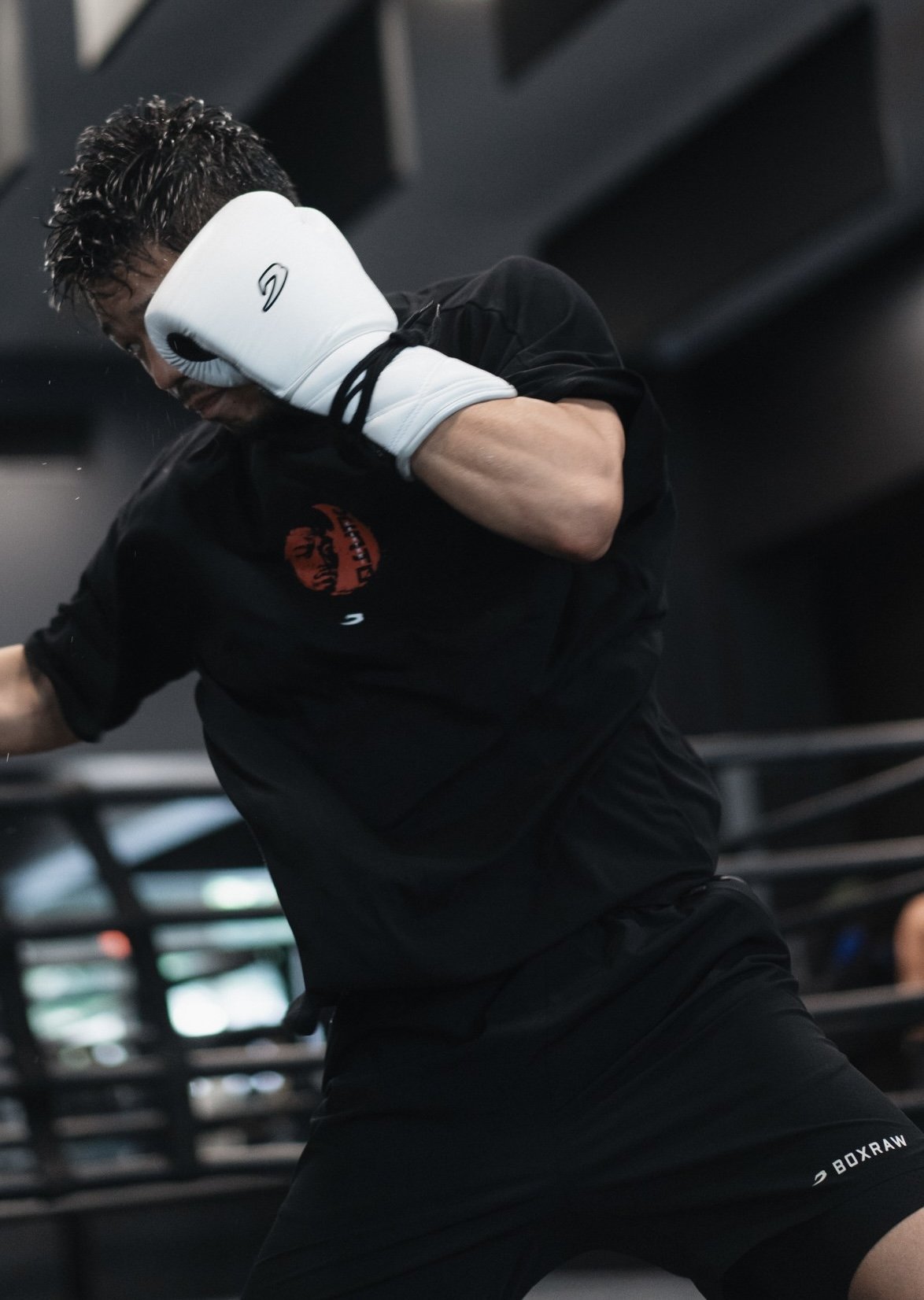Person wearing a black shirt and shorts practicing boxing with white boxing gloves in a gym.