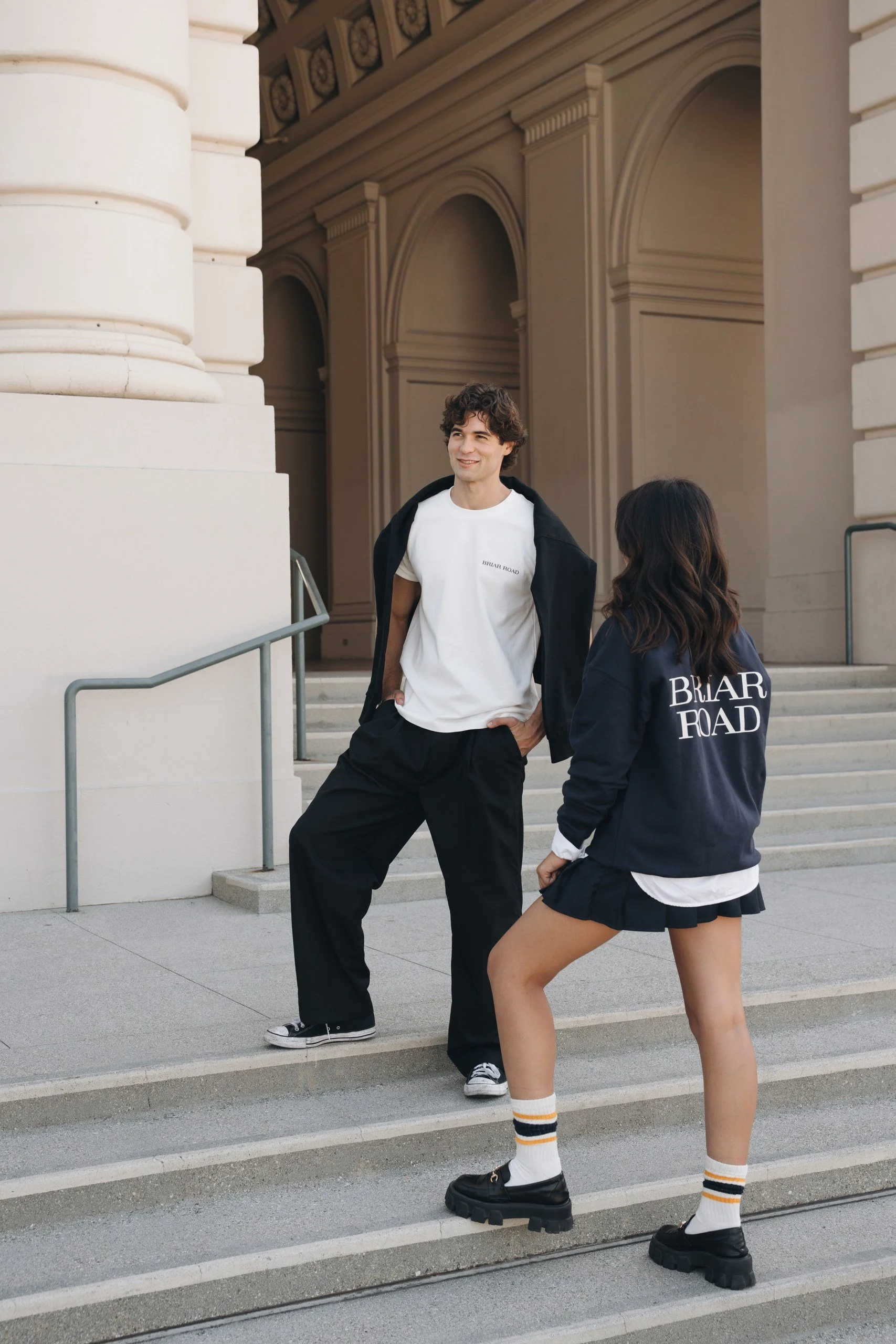 Two young people standing on steps outside a building with classical architecture. The man is wearing a white t-shirt, black jacket, and black pants, while the woman is dressed in a black jacket, white shirt, a black skirt, striped socks, and chunky 