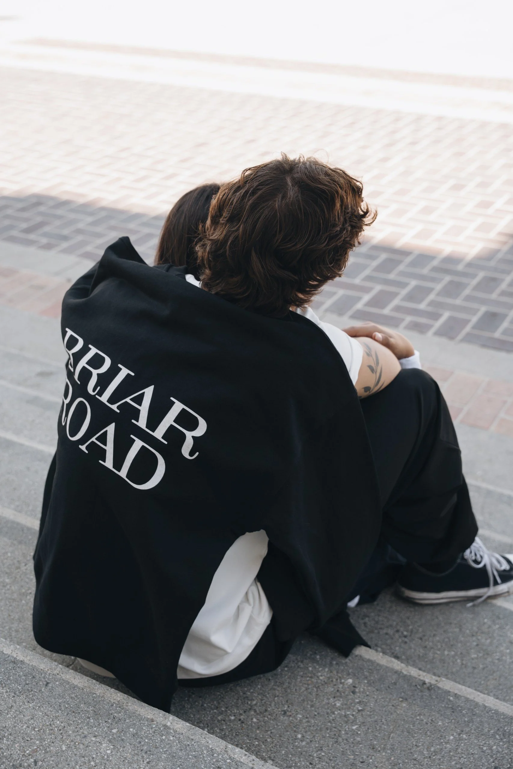 Two people sit on the ground outdoors, embracing. One person wears a black jacket with white text on the back, and the other wears a white garment. They are sitting on a paved surface with a brick1011 pattern. The scene appears emotional or comfortin