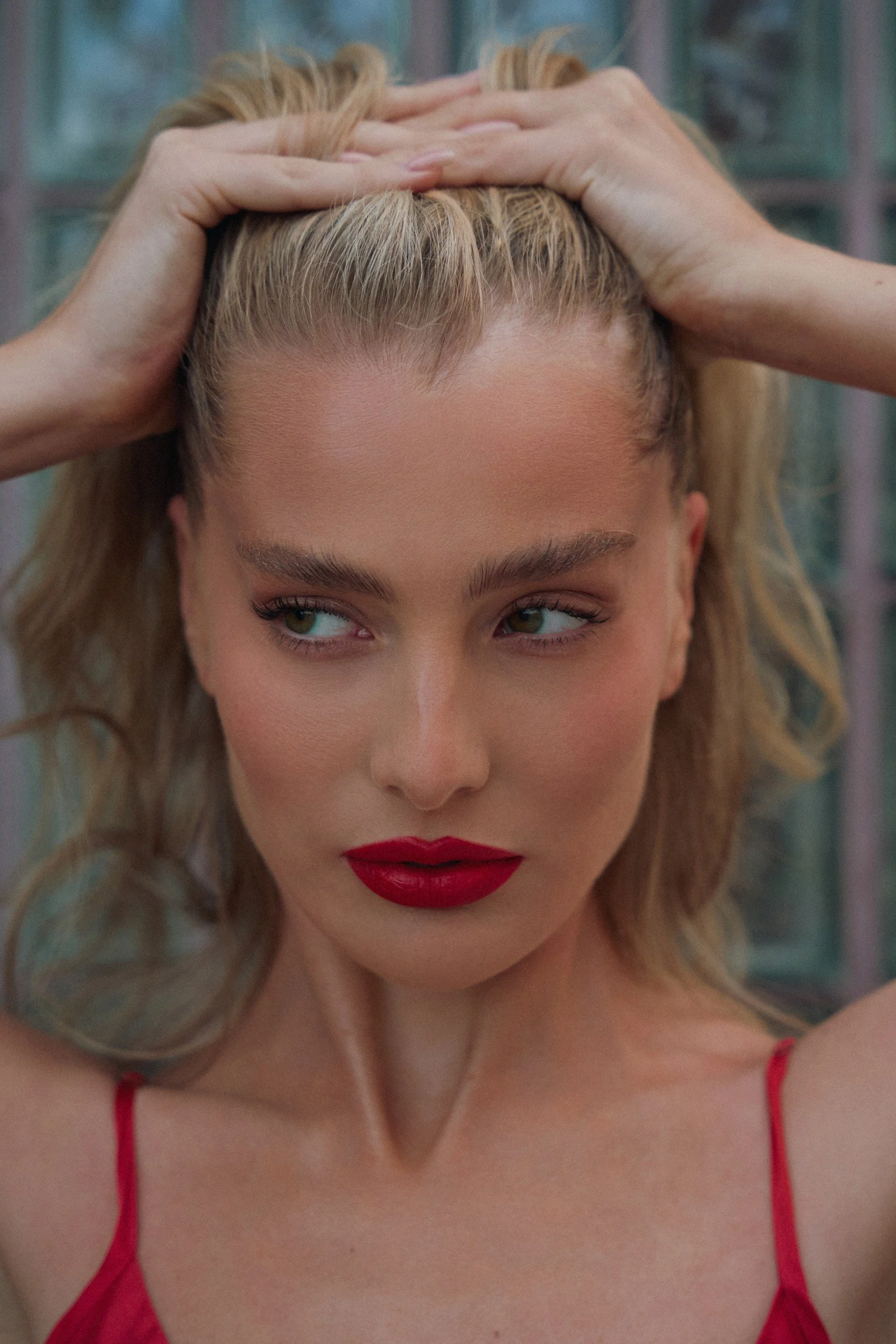 Close-up of a woman with blonde hair, red lipstick, wearing a red spaghetti strap top, with her hands on her head, looking down with a focused expression.