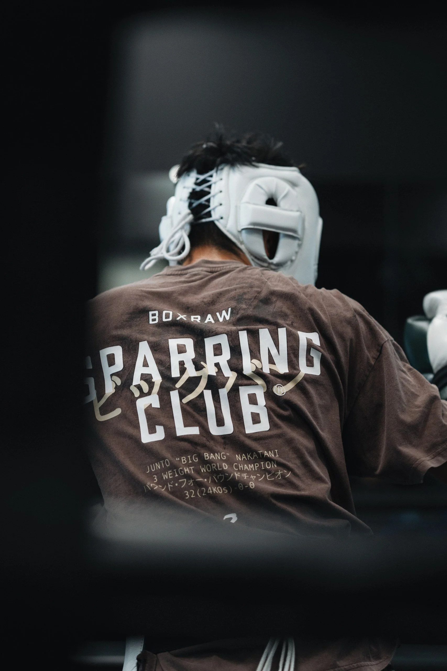 A person wearing a tan t-shirt with the words "Box & Raw" and "Parring Club" printed on the back, as well as Japanese text and mentions of a three-time world champion. The person is wearing a white boxing headgear and appears to be at a gym or traini