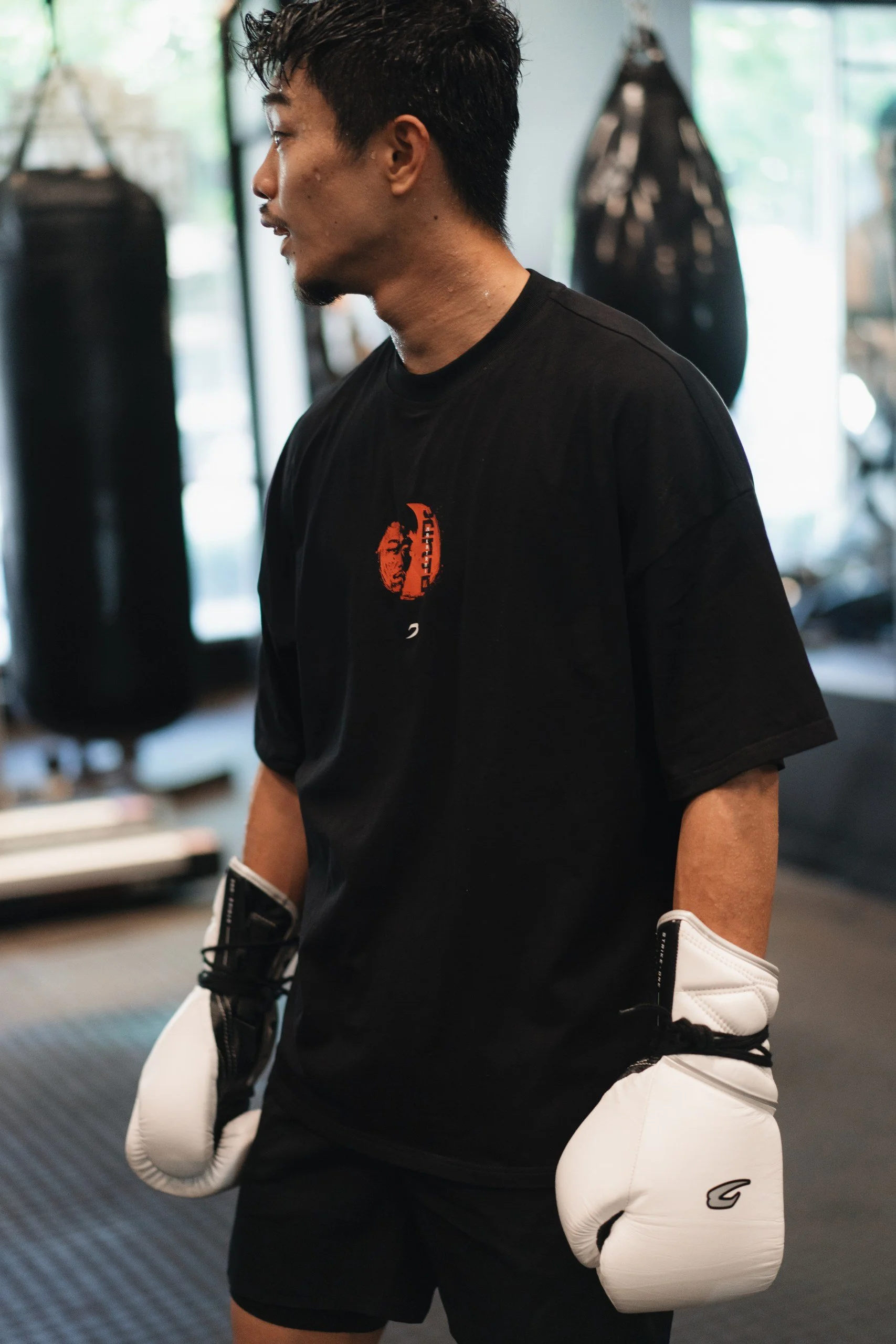 A person wearing boxing gloves, black T-shirt with red logo, and black shorts, standing in a gym with punching bags in the background.