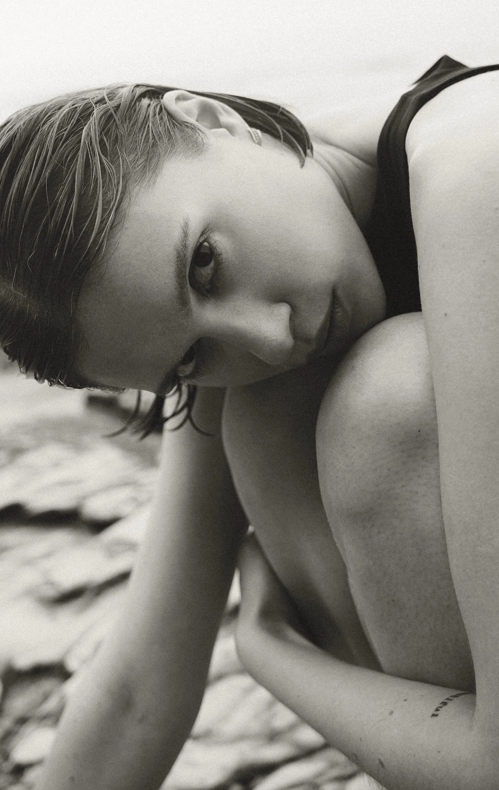 Close-up black and white photo of a woman with short hair lying on her side, partially covered by a blanket, looking towards the camera.