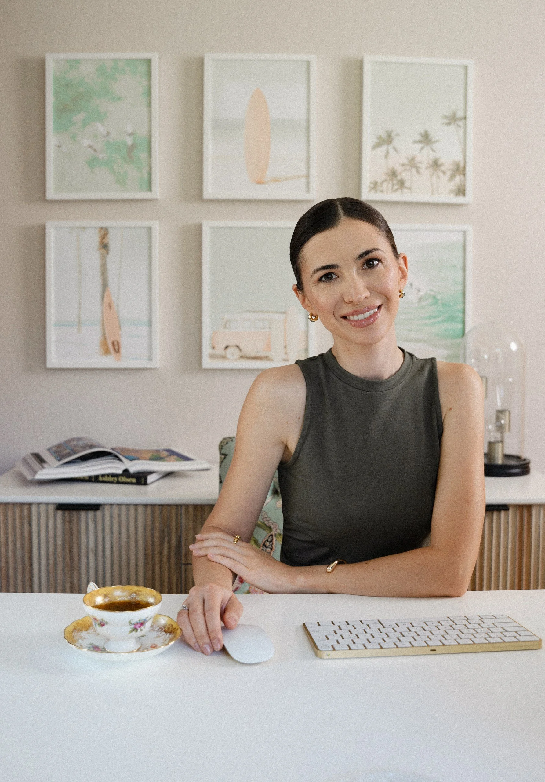 A woman with dark hair smiling at the camera, sitting at a white desk with a floral teacup and saucer, a computer mouse, and a keyboard. In the background, there is a wall with six framed art prints and a side table with books, a lamp, and decorative