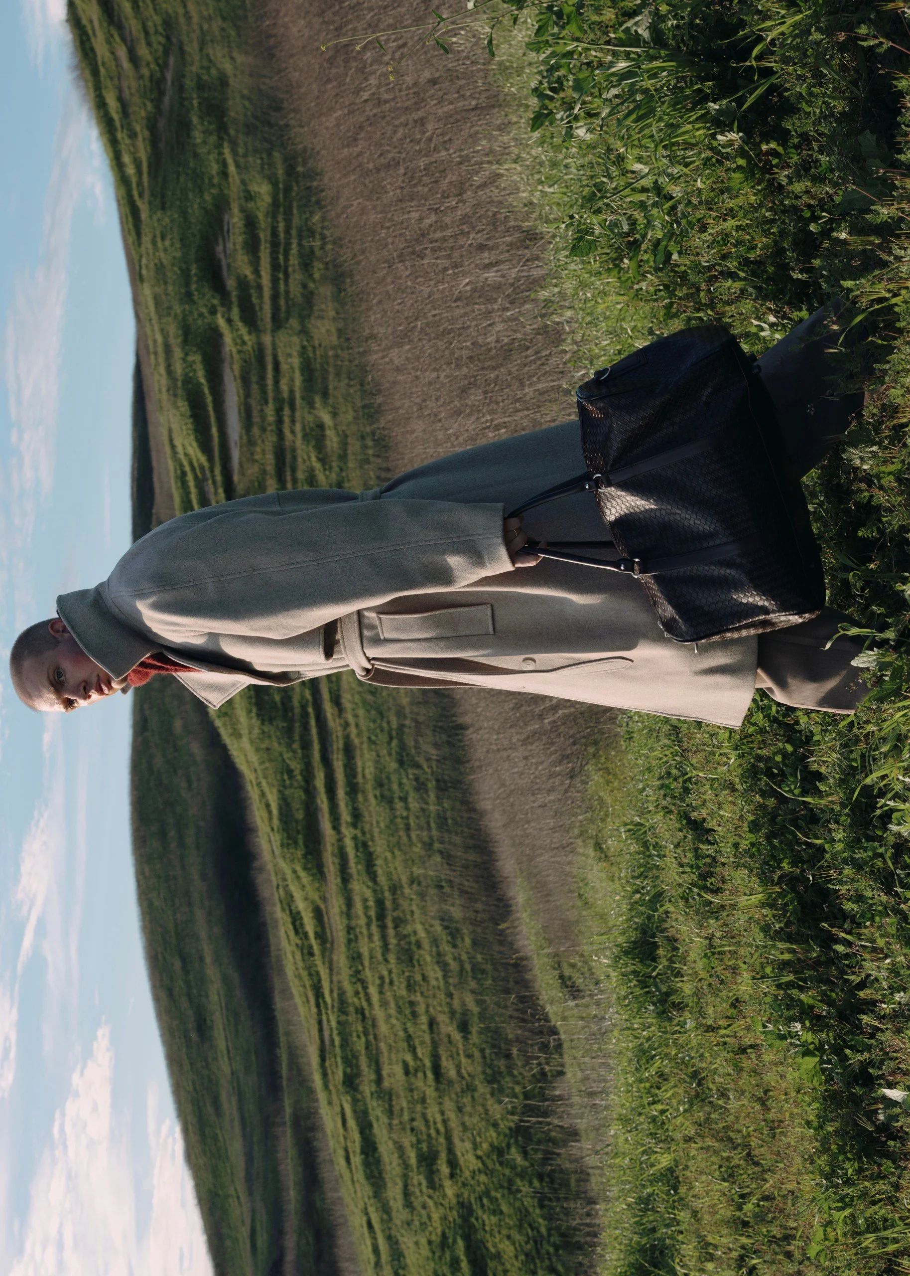 Person standing in a field of tall grass and plants, holding a black handbag, with a backdrop of greenery and blue sky.
