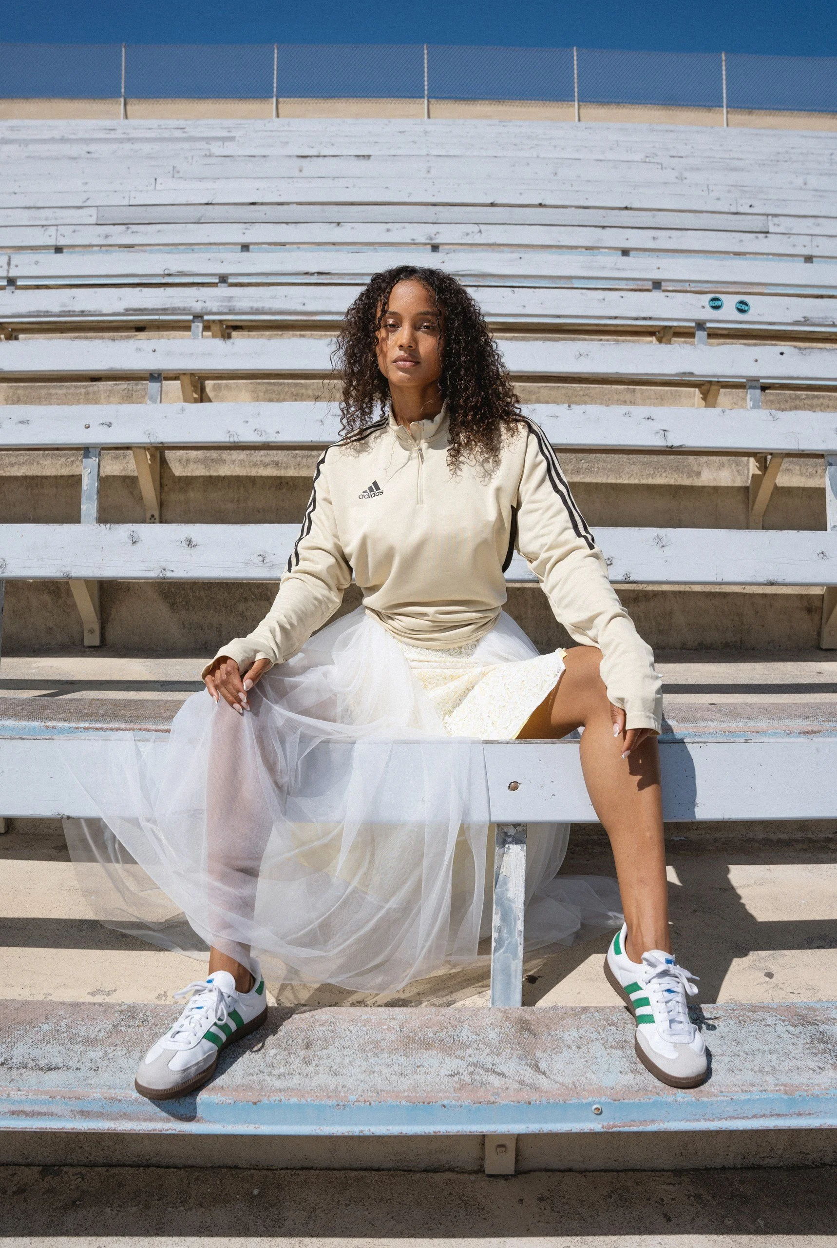A young woman with curly hair sitting on bleachers at an outdoor sports venue, wearing a beige Adidas sports jacket, a white skirt, and white sneakers with green accents, with a serious expression.