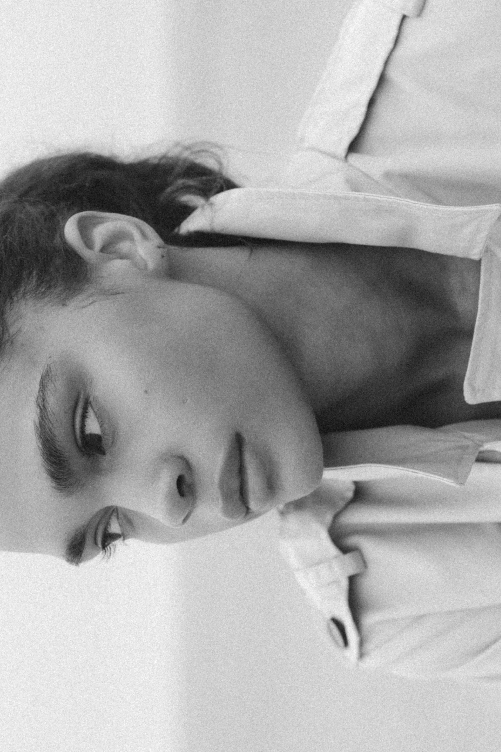 Close-up black and white photo of a woman with hair styled with soft curls, closed eyes, and slightly parted lips, lying on her side with her hand resting on her face.