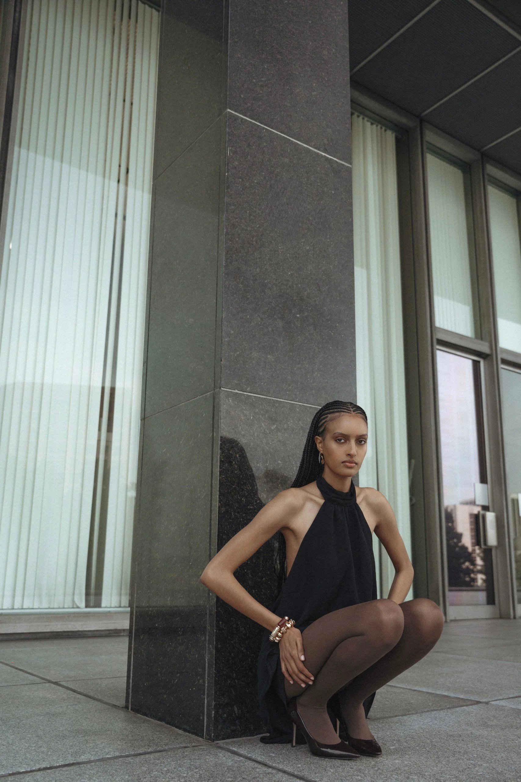 A woman in a black dress with a halter neck, black stockings, and high heels crouches against a building corner with large windows and vertical blinds. She has braided hair, wears bracelets, and has a composed expression.