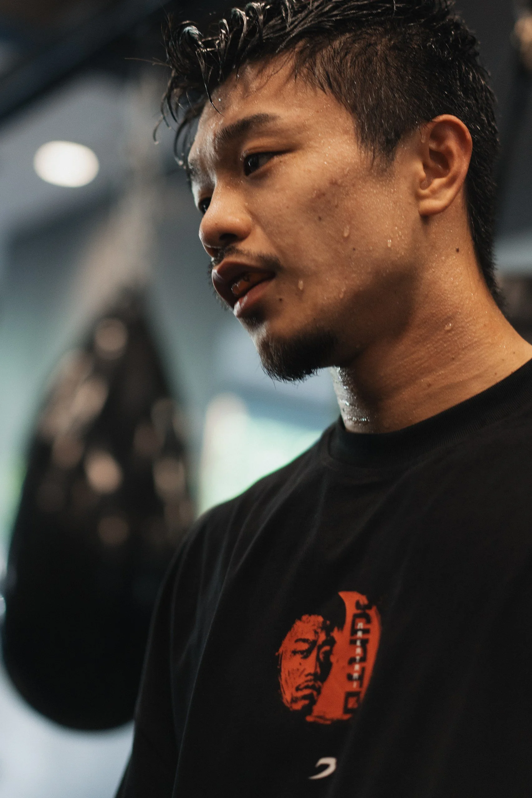 A young man with black, wet hair is in a gym, lifting weights. He is wearing a black T-shirt with an orange graphic. Sweat is visible on his face and neck.
