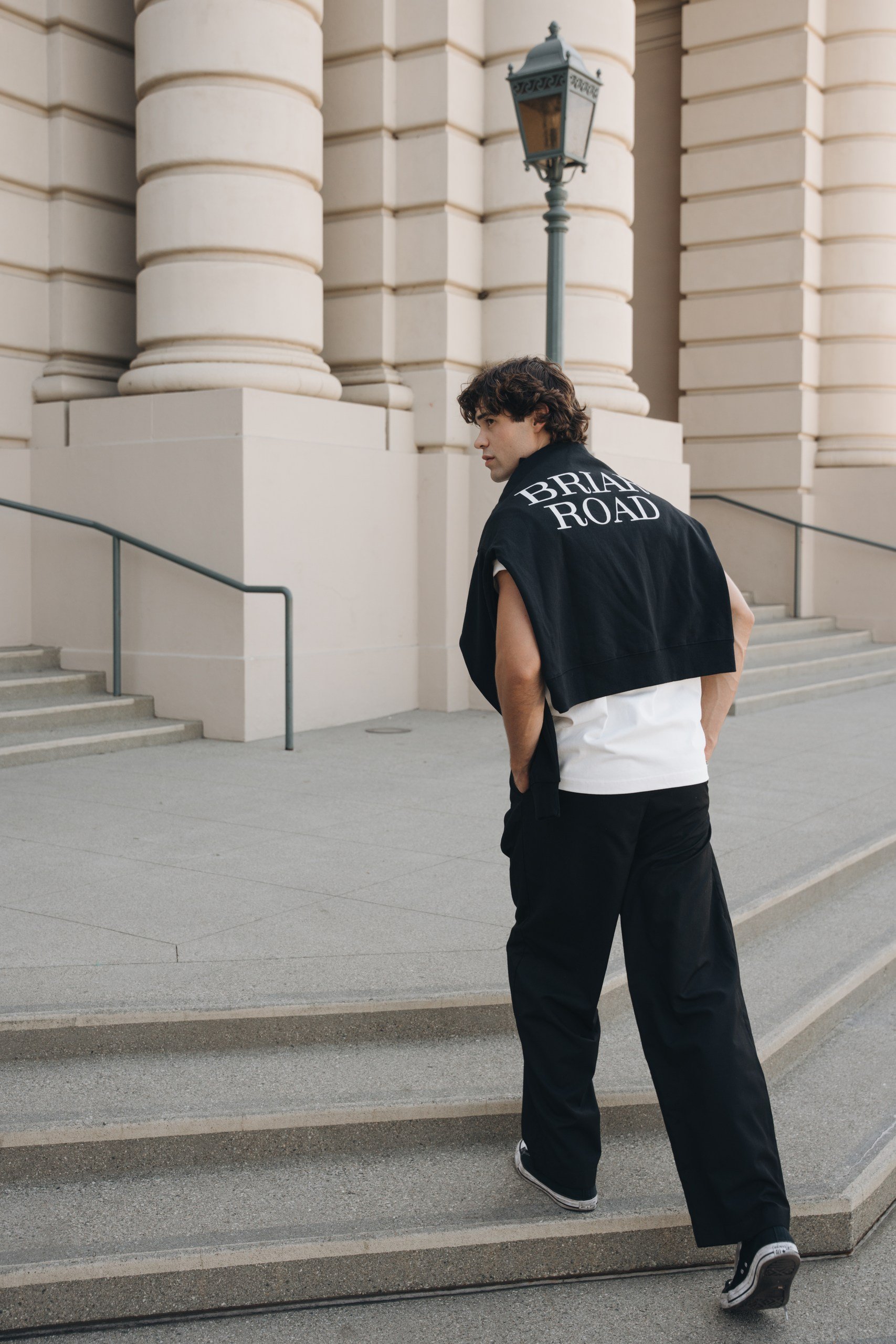 A young man with curly dark hair wearing a black jacket with white text on the back, walking up stairs outside a large building with architectural columns and a vintage-style lamp post.