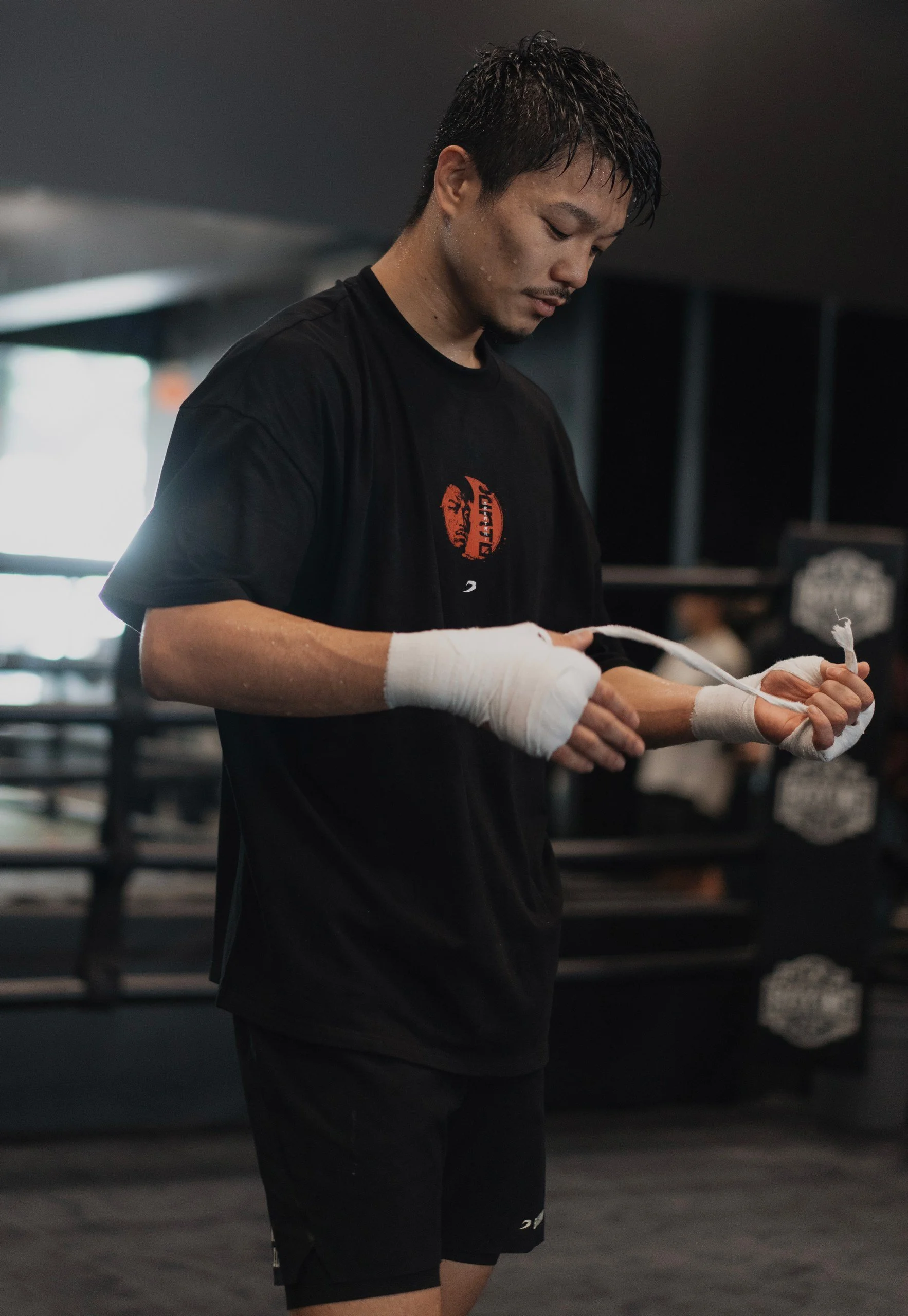 A young man with short dark hair and a goatee, wrapped hands, and wearing a black T-shirt and shorts, looks down while preparing in a gym or boxing training facility.