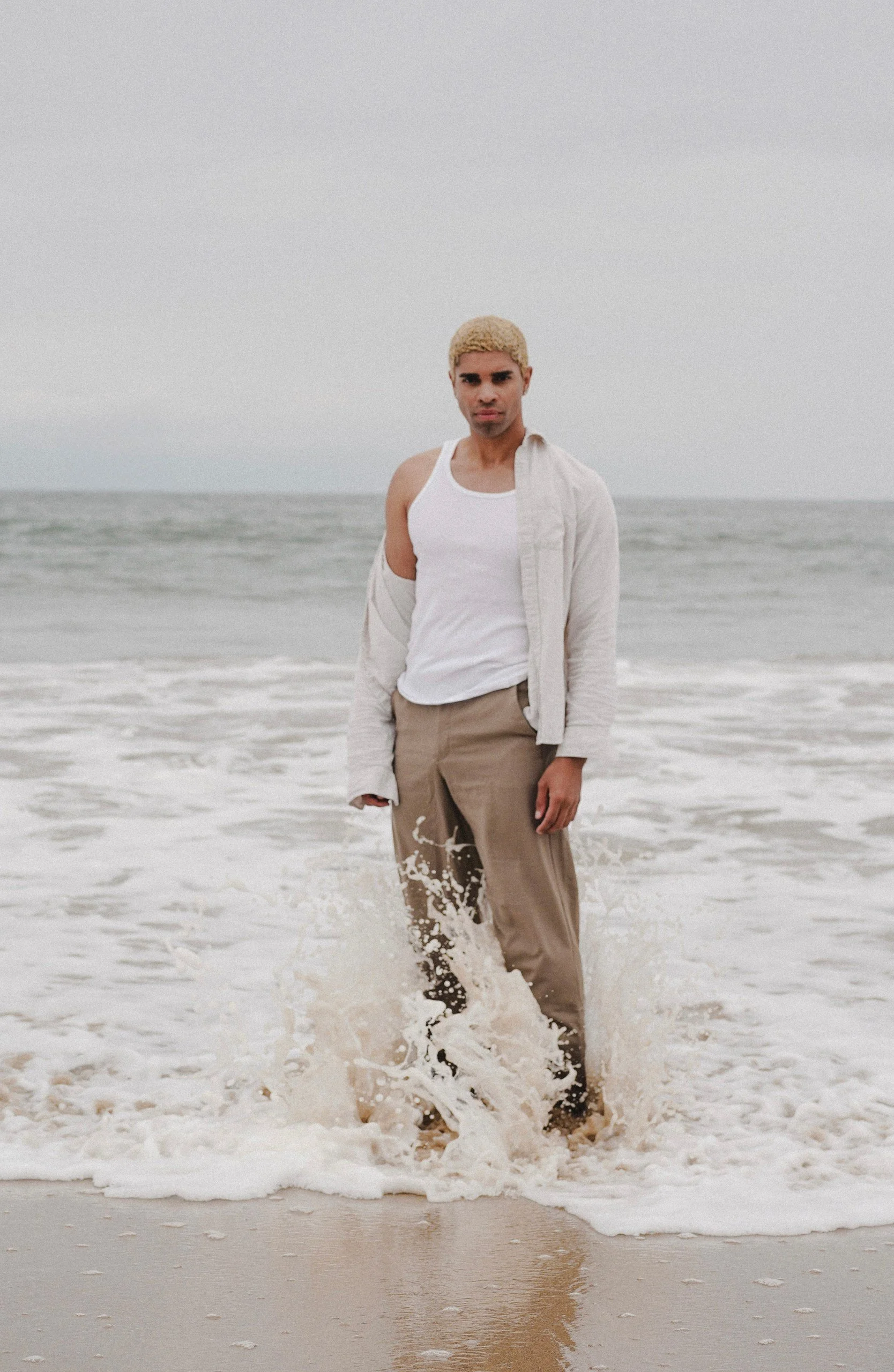 A man with bleached blonde hair standing in the ocean with waves around his legs, wearing a white tank top and beige pants, under an overcast sky.