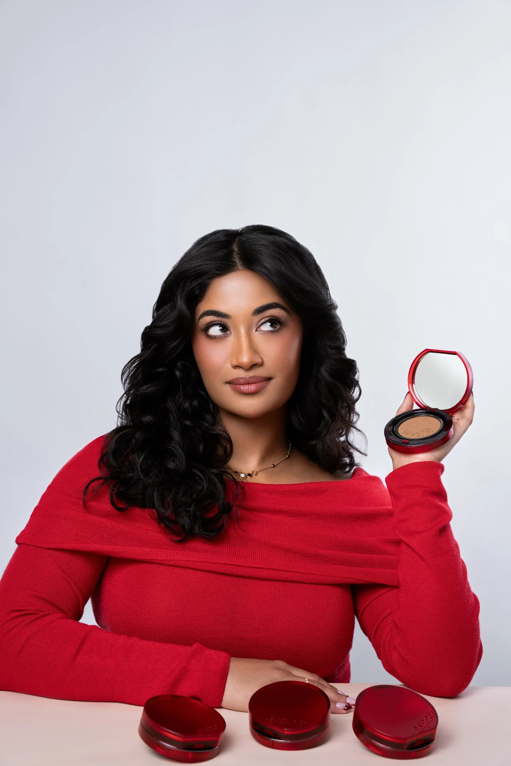 Woman with long dark hair and a red shirt, holding an open compact with beige makeup, sitting at a white table with three closed red compacts in front of her against a gray background.