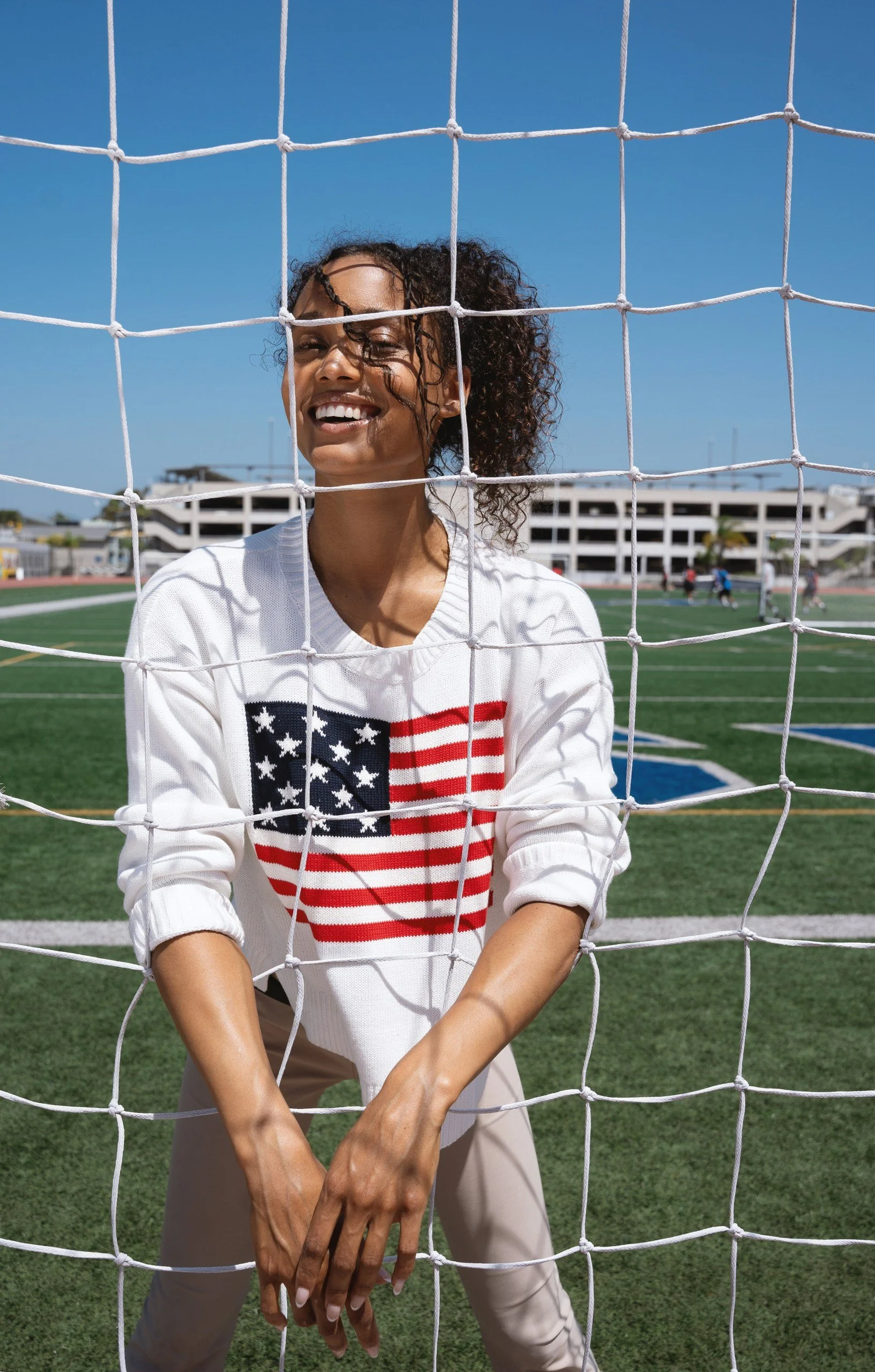 A woman stands behind a soccer net on a field, smiling at the camera. She is wearing a white shirt with an American flag design and beige pants, with her hands resting in front of her. There are people and a building in the background under a clear b