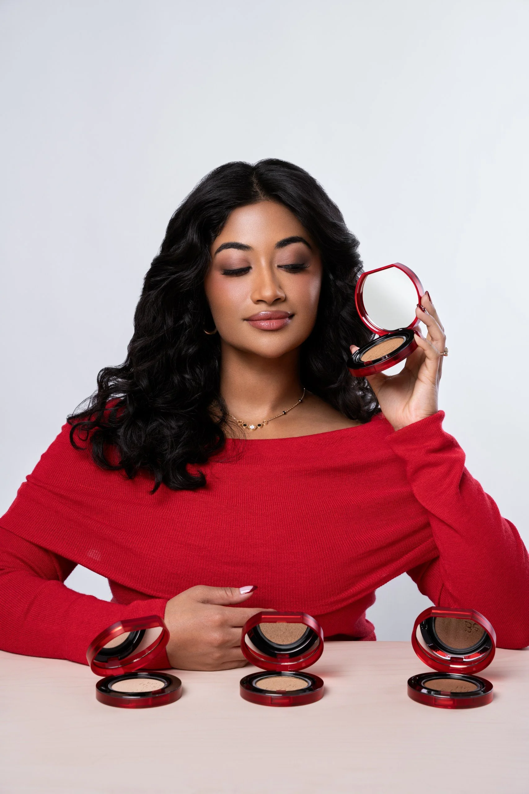 Woman in red top holding a red compact makeup mirror with makeup products on table.