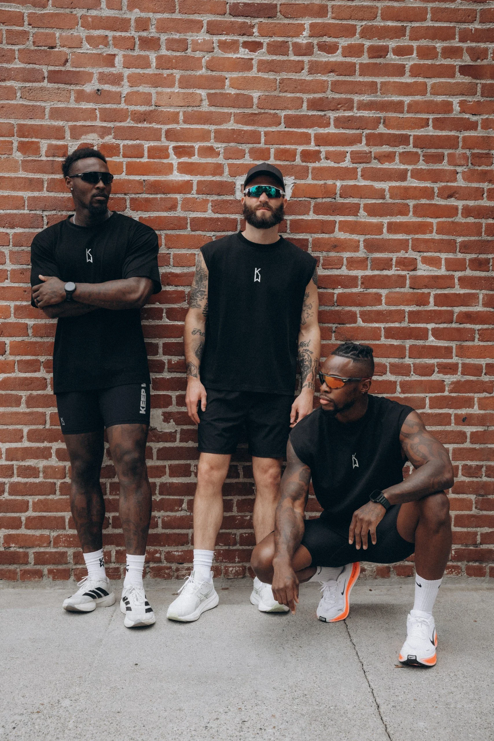 Four men wearing black athletic clothing and white sneakers are posing against a red brick wall. Three are standing, one is crouching down, all wearing sunglasses, with visible tattoos on their arms, and one with a cap.