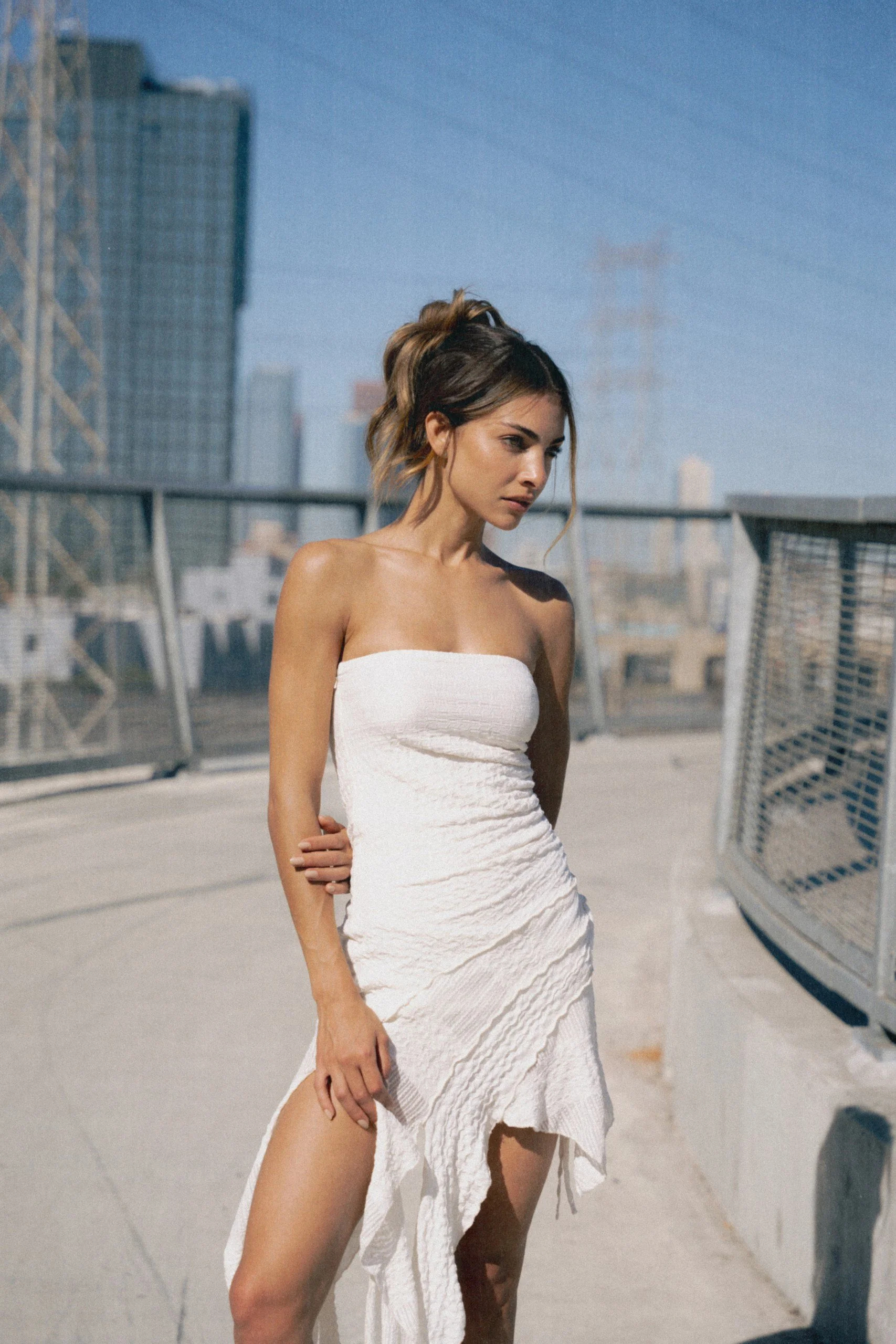 A young woman in a strapless white dress standing outdoors on a rooftop with city skyline in the background.