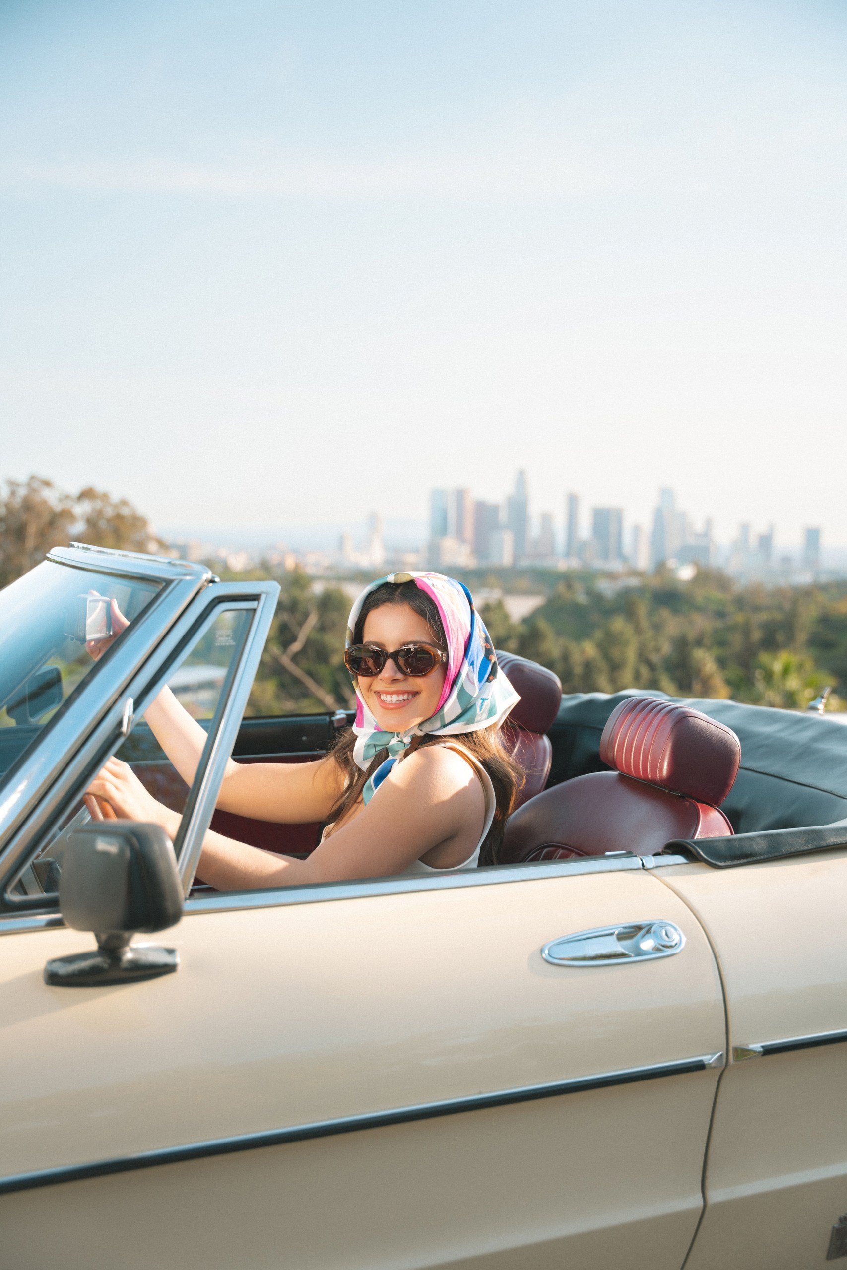 A woman wearing sunglasses and a colorful headscarf driving a cream-colored convertible car with red seats, with a city skyline visible in the background.