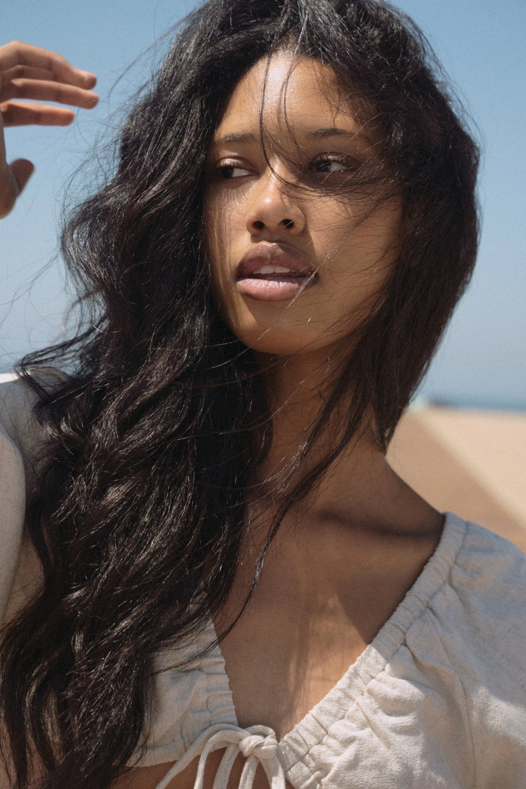 A woman with long dark wavy hair looking to her right, outdoors with a blue sky background, wearing a light-colored top with a gathered front and a bow.