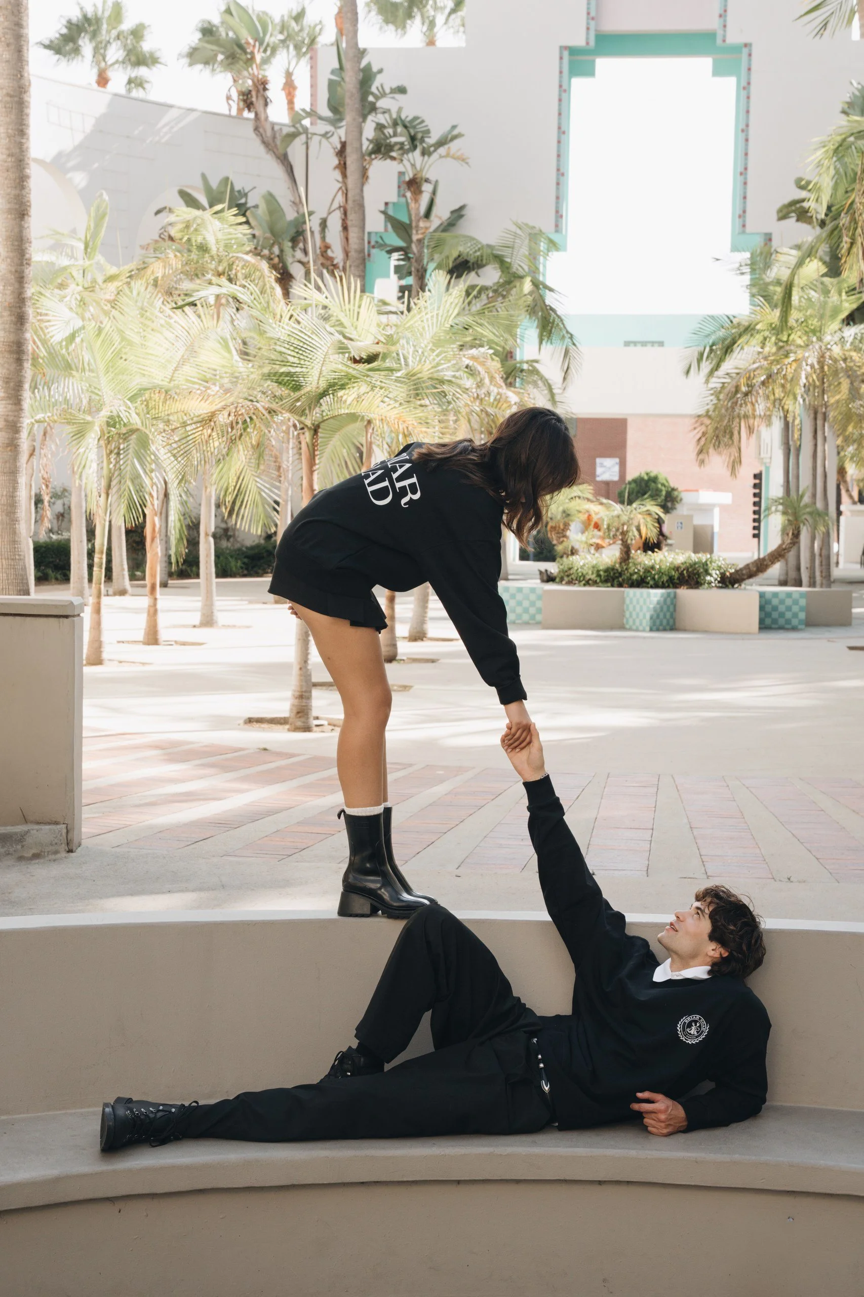 A woman with long dark hair in a black sweatshirt and tall black boots bending down to hold the hand of a man lying on the ground. The man has curly hair, wears a black jacket and pants, and is looking up at the woman. They are outdoors in a sunny ar