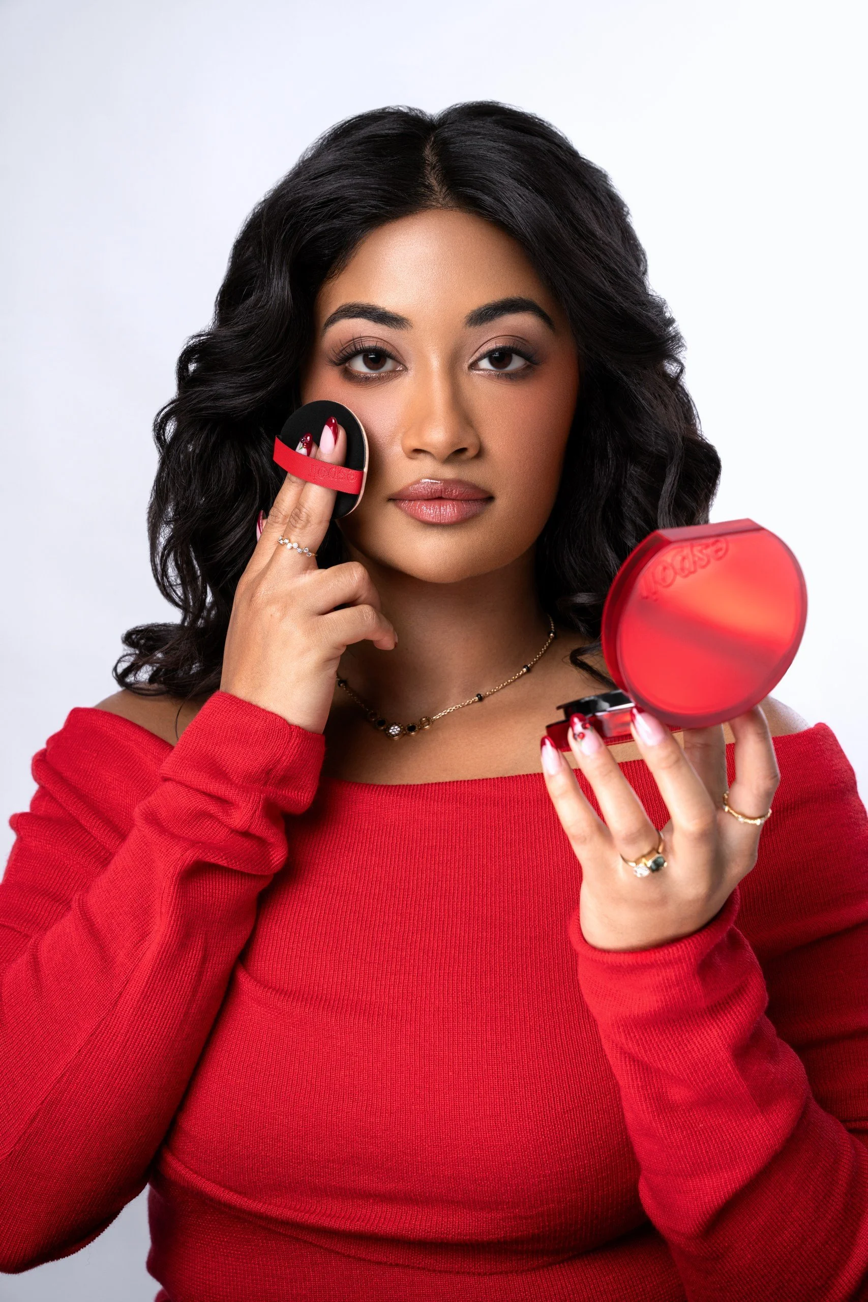 A woman with dark, curly hair applies makeup with a sponge near her cheek while looking into a mirror. She is wearing a red top and jewelry, and has a neutral background.