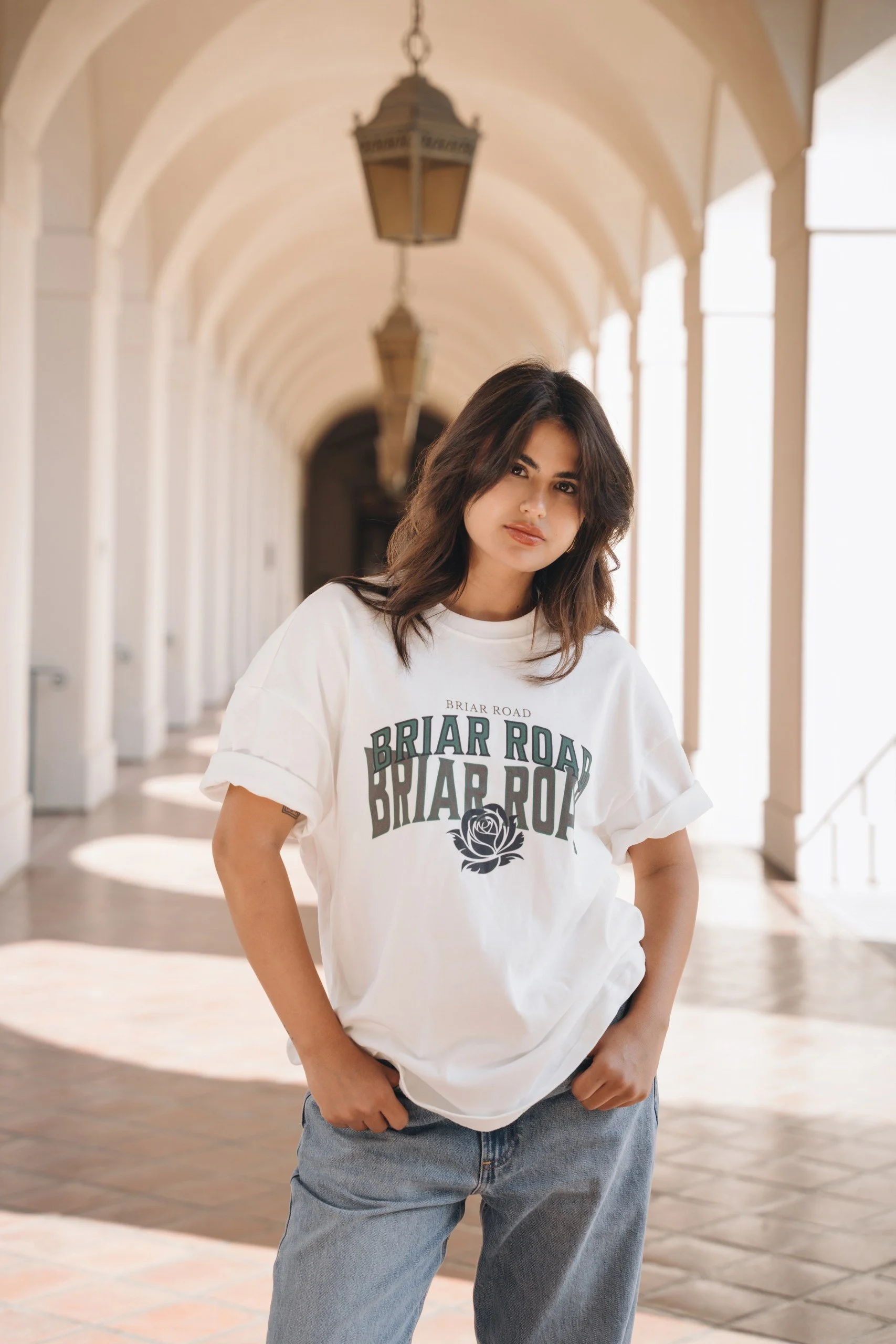 Young woman with shoulder-length dark hair, wearing a white t-shirt and jeans, standing in an arched corridor with hanging lanterns.