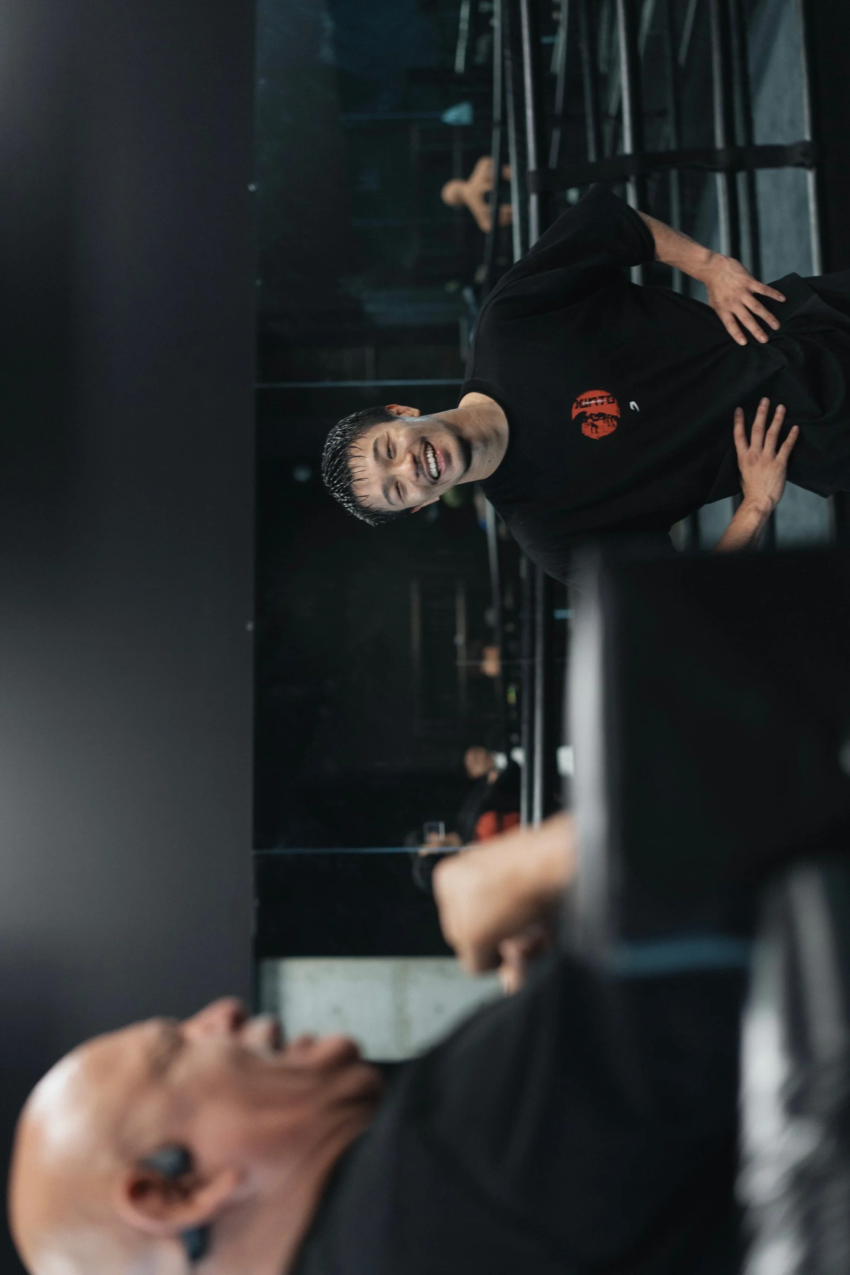 A young man in a black shirt with a red logo is smiling and posing with hands on his hips in an indoor setting, with other blurred people in the background.