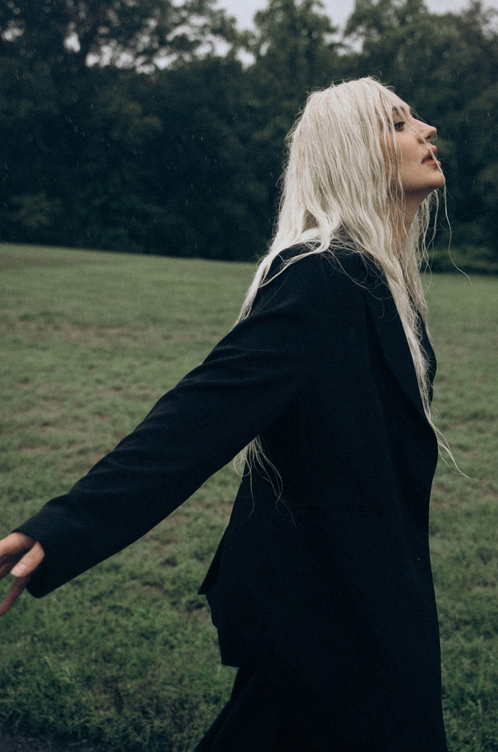 A woman with platinum blonde hair wearing a black outfit standing outdoors with arms outstretched in a grassy field during a rain shower.