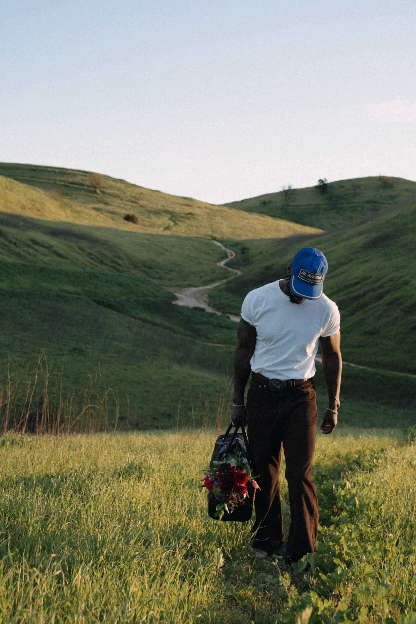 A man walking through a grassy field holding a bouquet of red and purple flowers in one hand and a camera in the other. He is wearing a white t-shirt, dark pants, and a blue baseball cap. The landscape features rolling green hills under a clear sky.